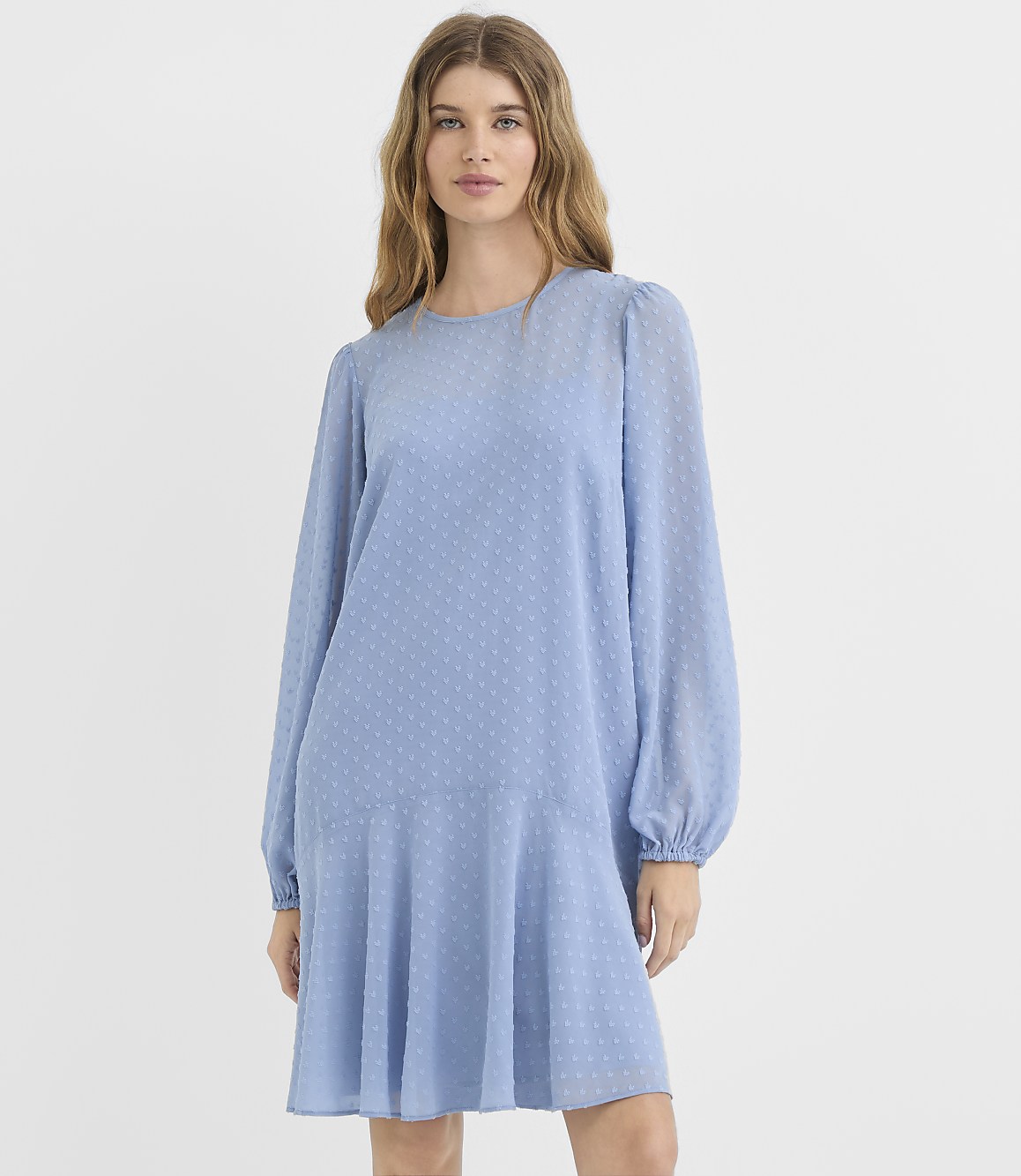 A woman in a light blue, textured dress with voluminous sleeves stands against a plain white background.