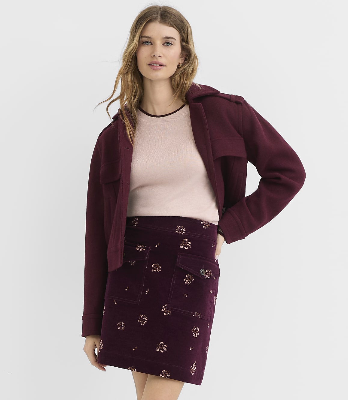 A woman in a burgundy jacket and floral skirt stands against a plain white background.