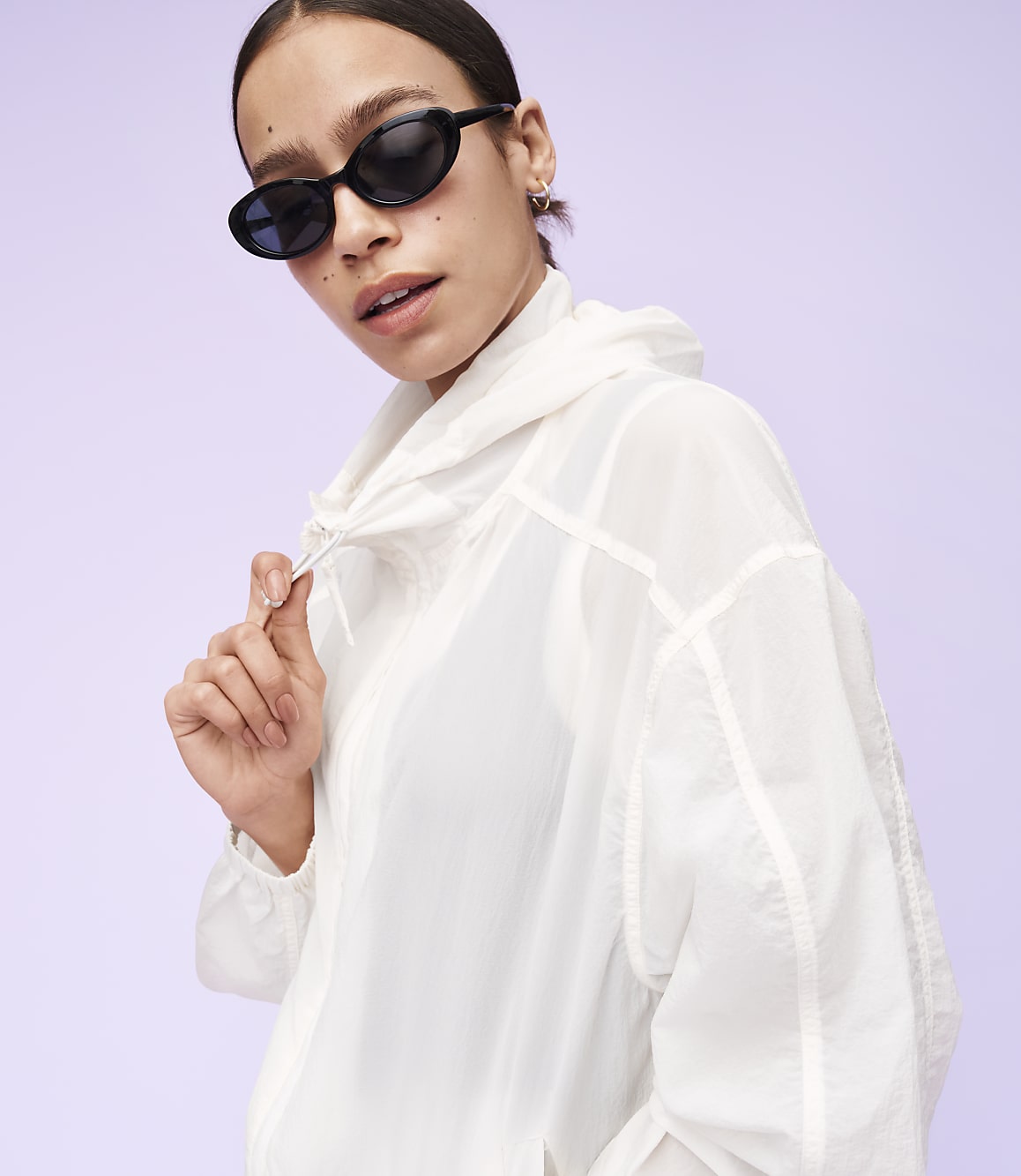 A woman in a white jacket and sunglasses poses against a light purple background.