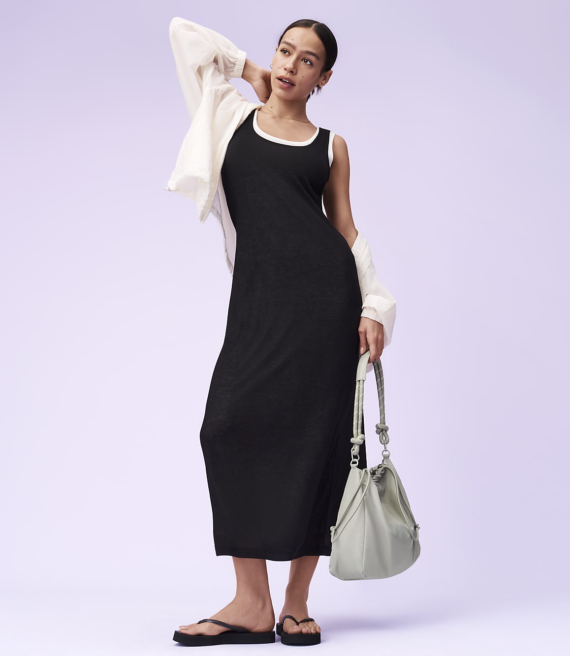A woman in a black dress and white cardigan stands with a light green purse against a lavender background.