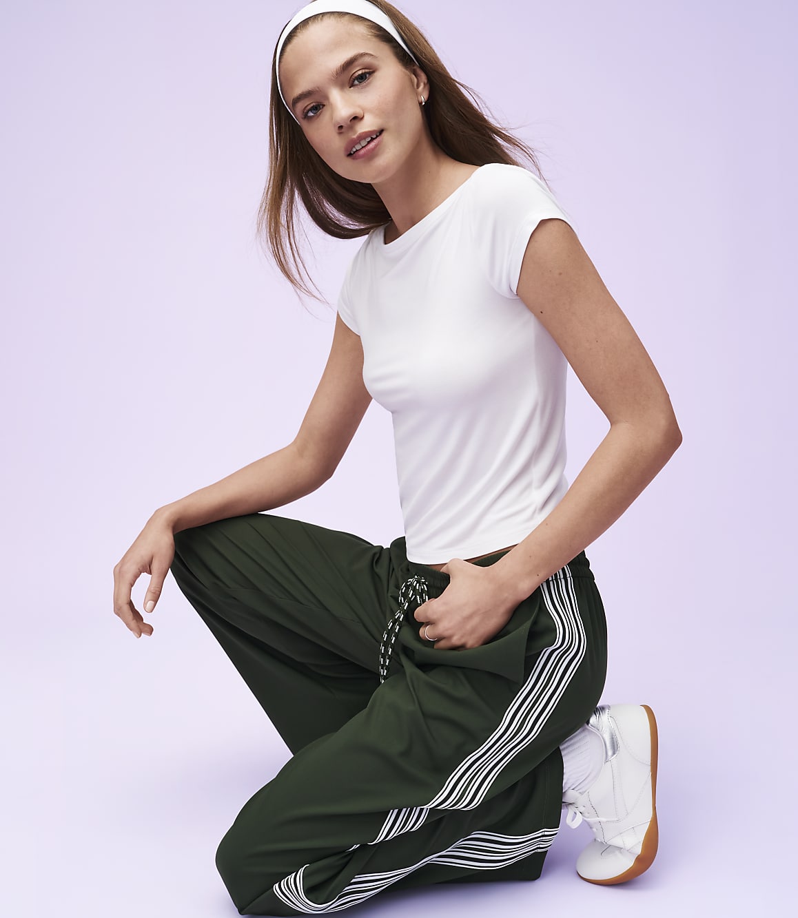 A young woman in a white t-shirt and green track pants with white stripes kneels against a soft purple background.