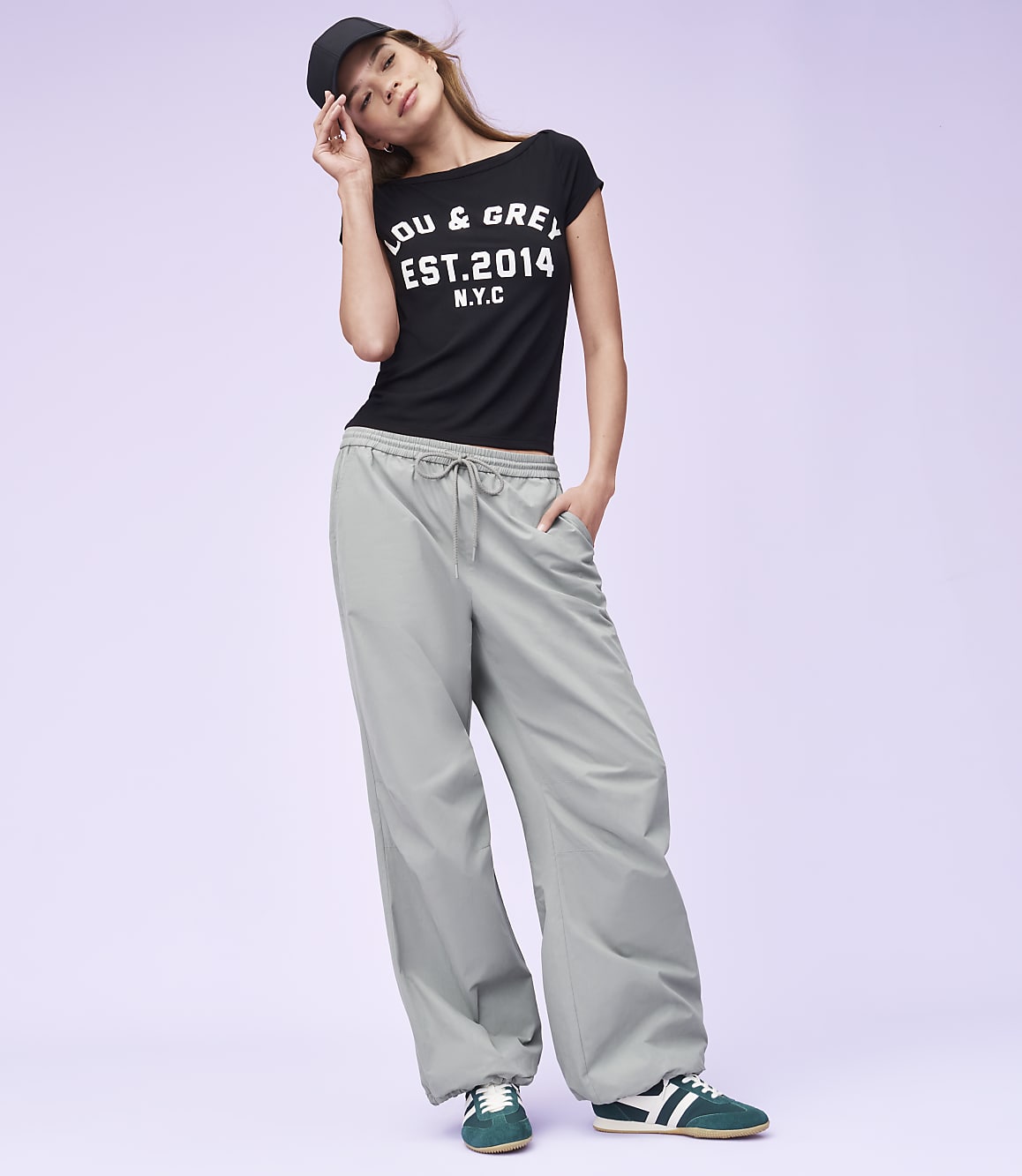 A woman in a black t-shirt and grey pants poses against a plain purple background.
