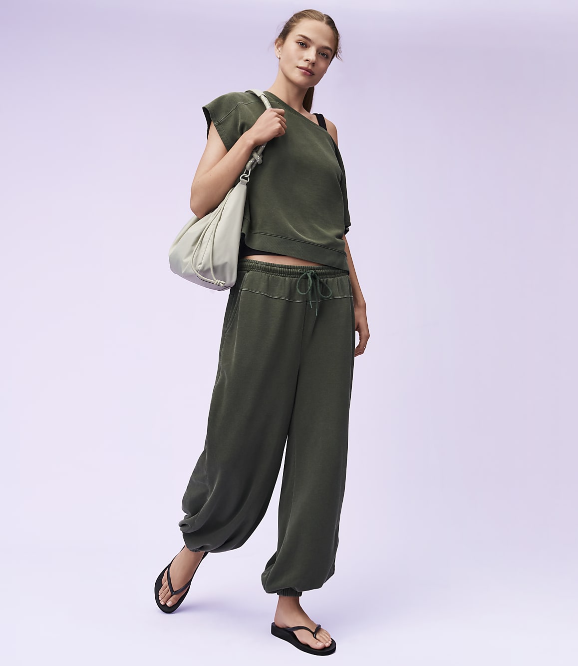 A woman in a green outfit and flip-flops stands with a bag over her shoulder against a light purple background.