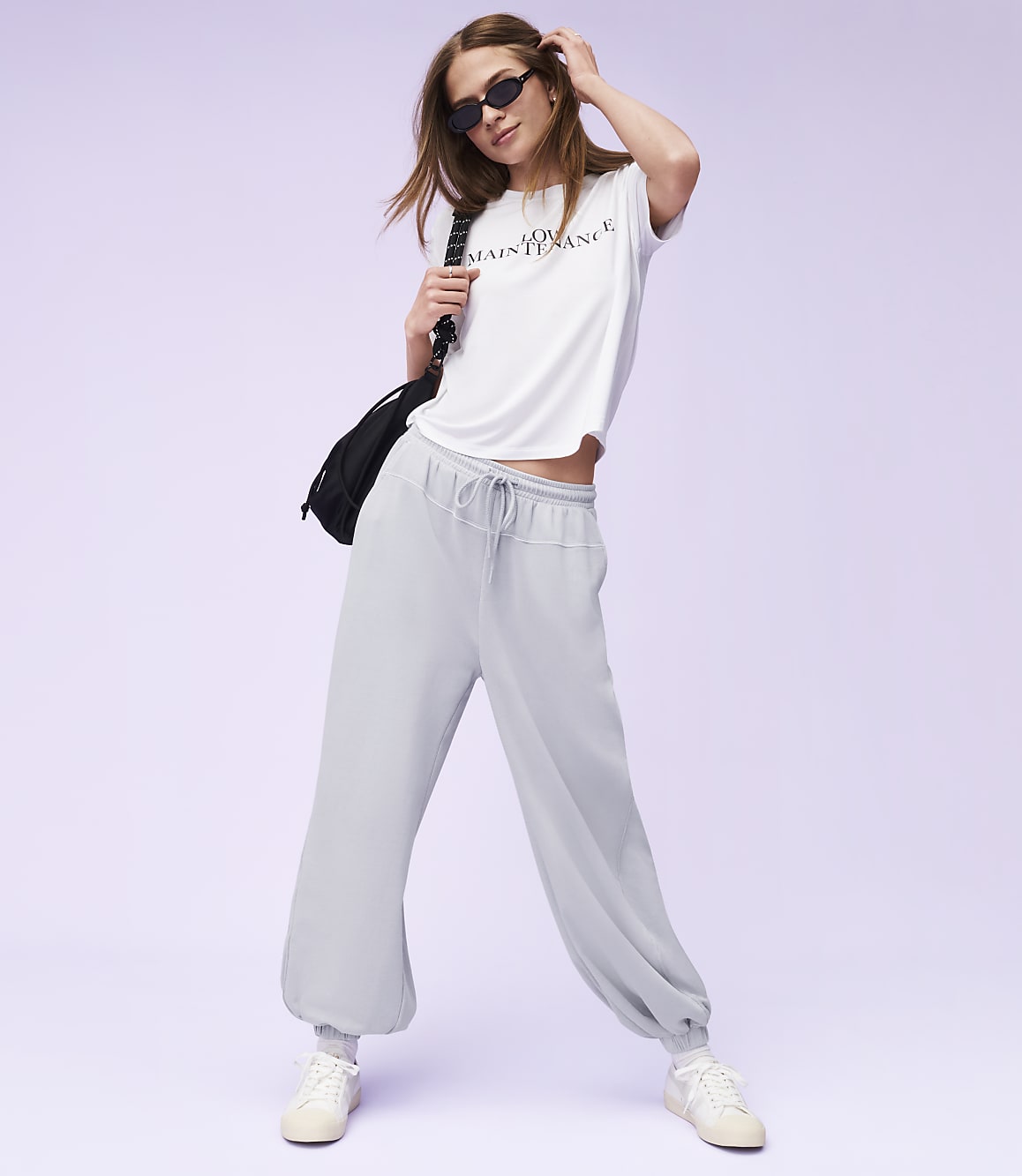 A woman in a white t-shirt and light blue sweatpants poses against a soft purple background.