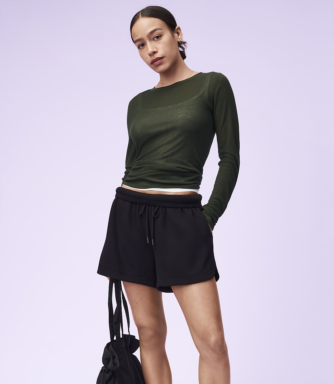 A woman in a green long-sleeved shirt and black shorts stands against a light purple background, holding a black bag.
