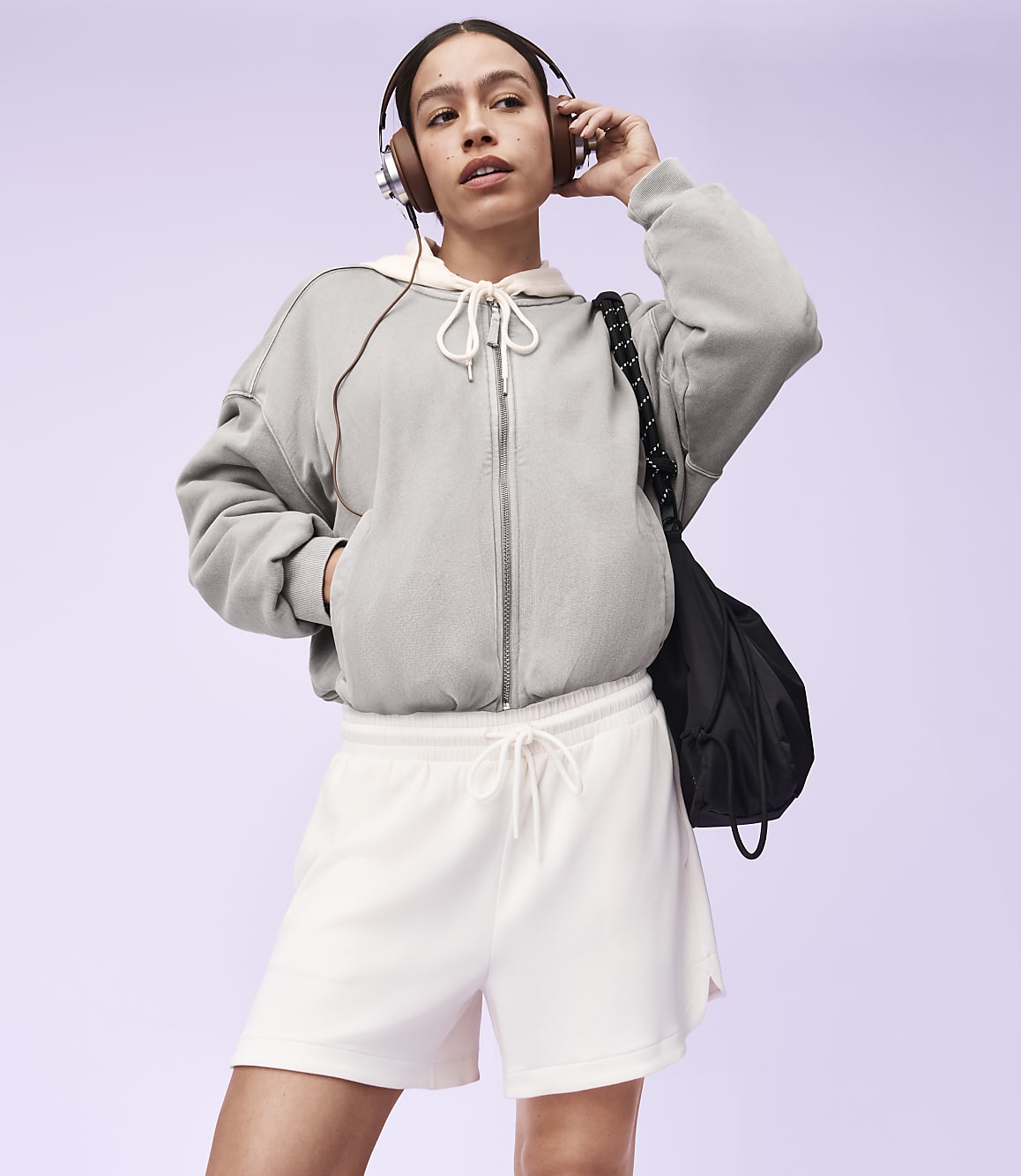A young woman in a grey zip-up hoodie and white shorts stands with headphones on, a black bag slung over her shoulder, against a soft purple background.