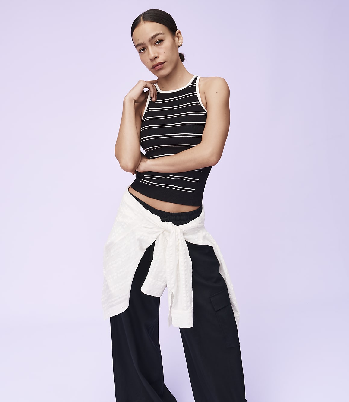 A woman in a striped tank top and black pants stands against a plain purple background, with a white sweater tied around her waist.