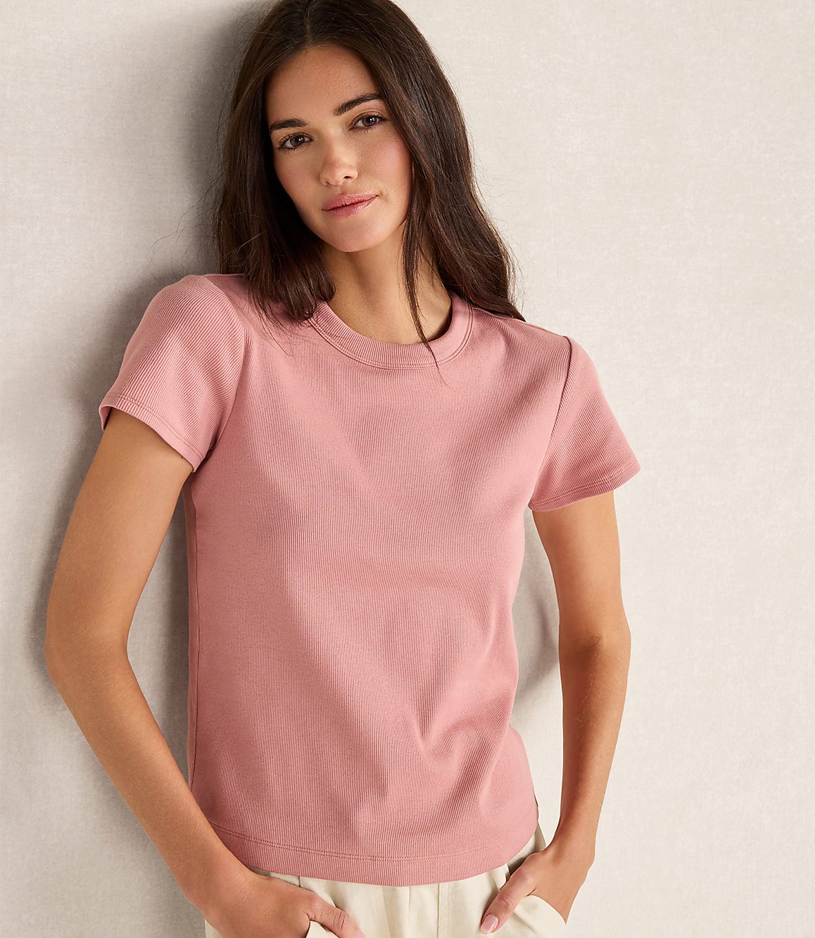 A woman in a pink ribbed t-shirt stands against a textured, light-colored wall.