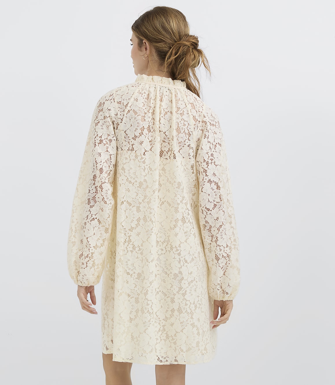 A woman in a cream lace dress stands against a plain white background.
