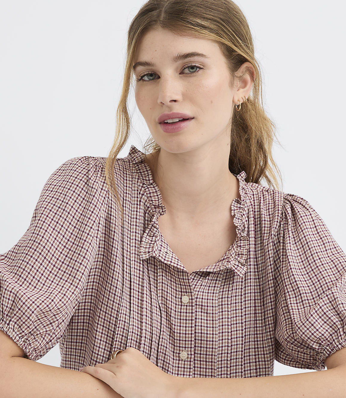 A young woman with blonde hair pulled back wears a pink and white checkered blouse with a ruffled collar, against a plain white background.