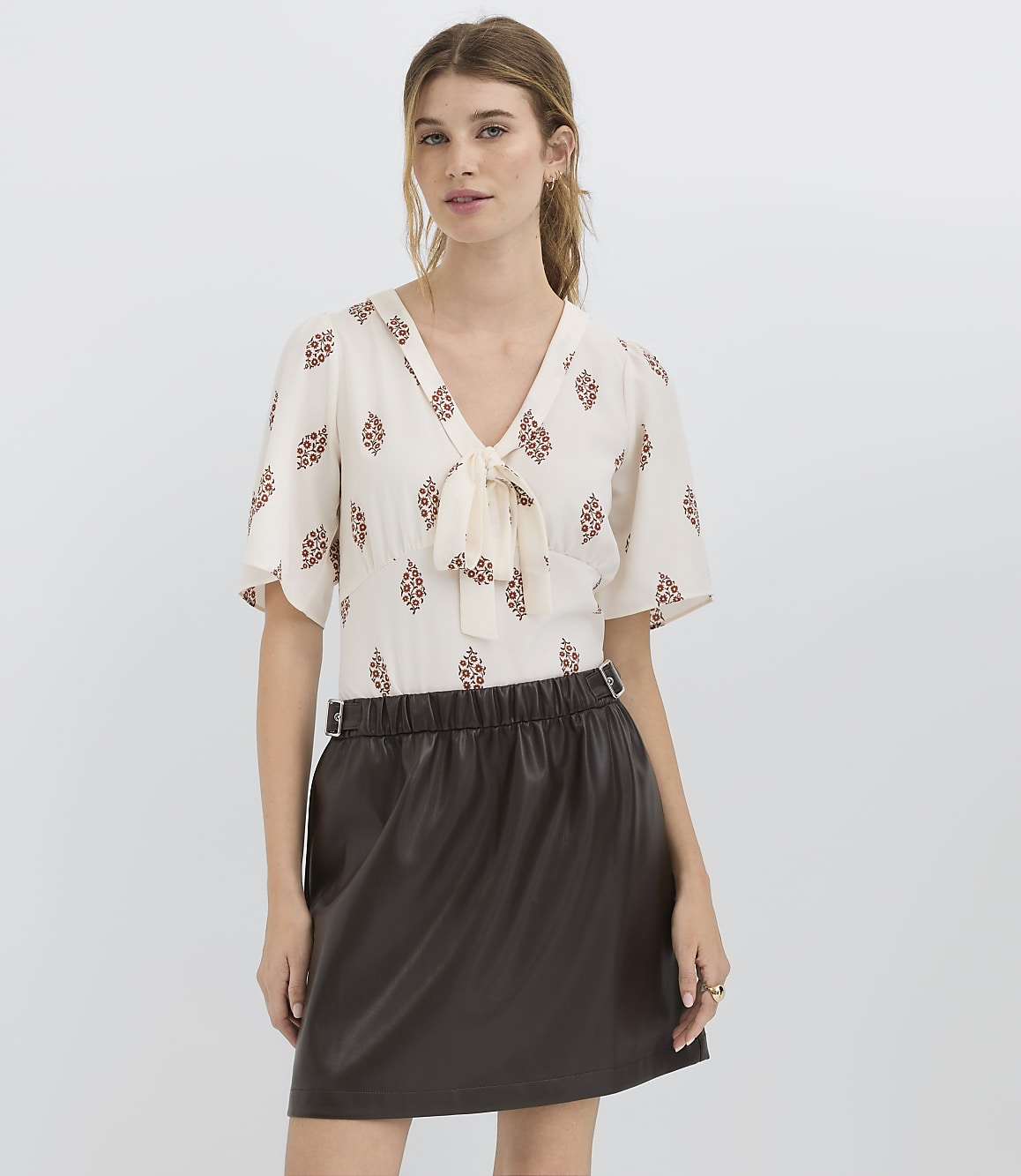 A woman in a patterned blouse and a dark skirt stands against a plain white background.