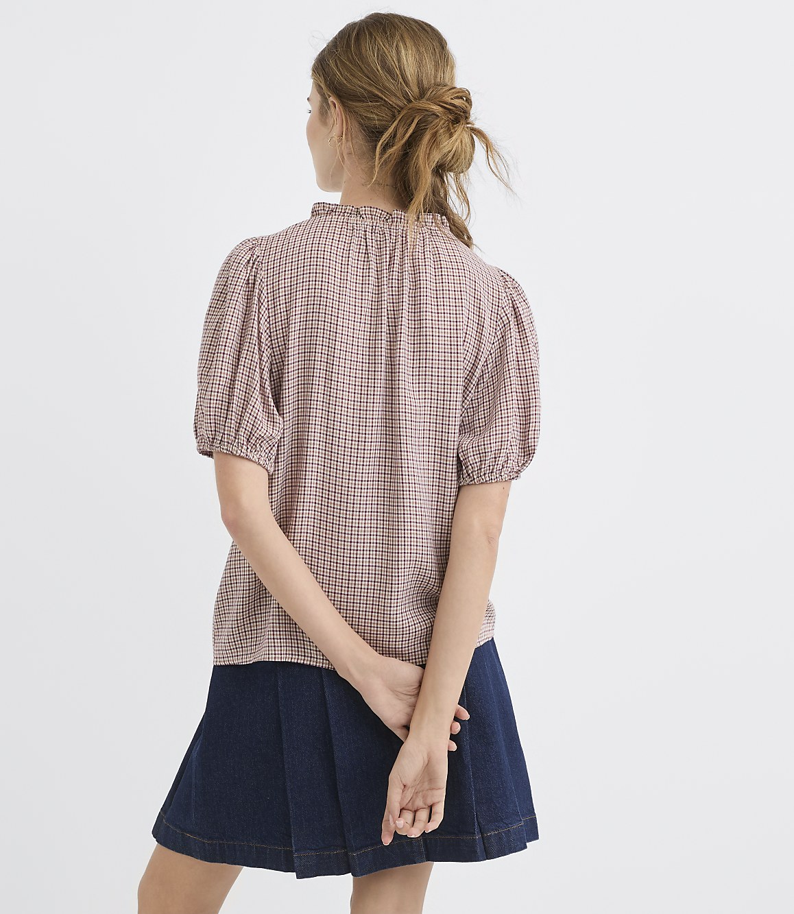 A woman in a patterned blouse and denim skirt stands with her back to the camera against a plain white background.