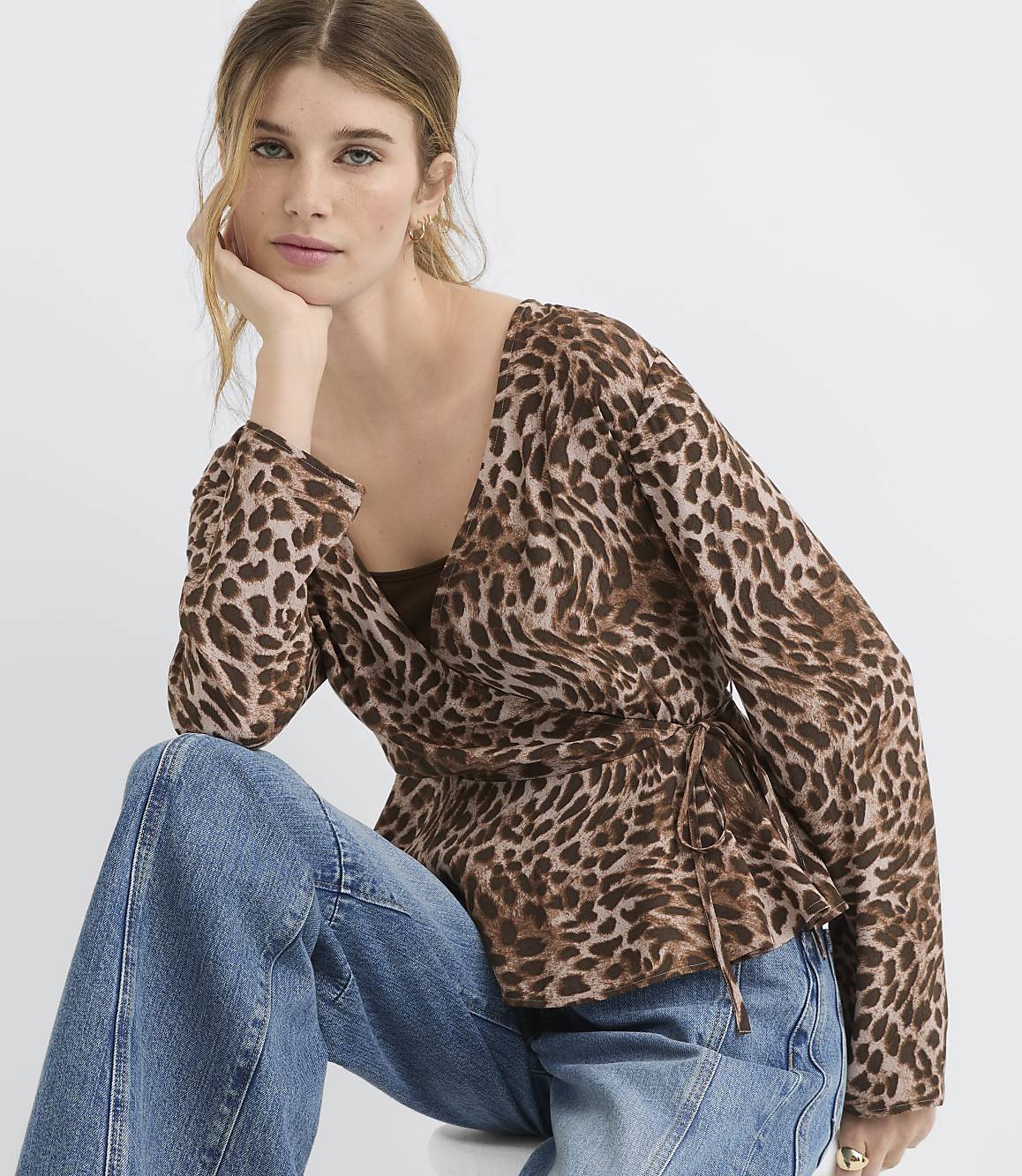 A woman in a leopard-print wrap top and blue jeans poses against a plain white background.