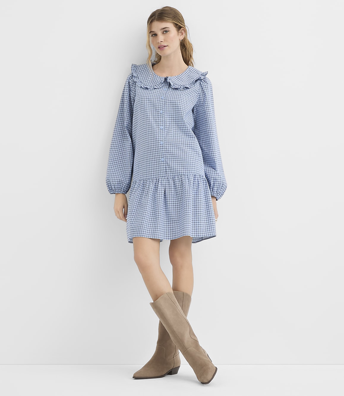 A young woman in a blue gingham dress and taupe boots stands against a plain white background.