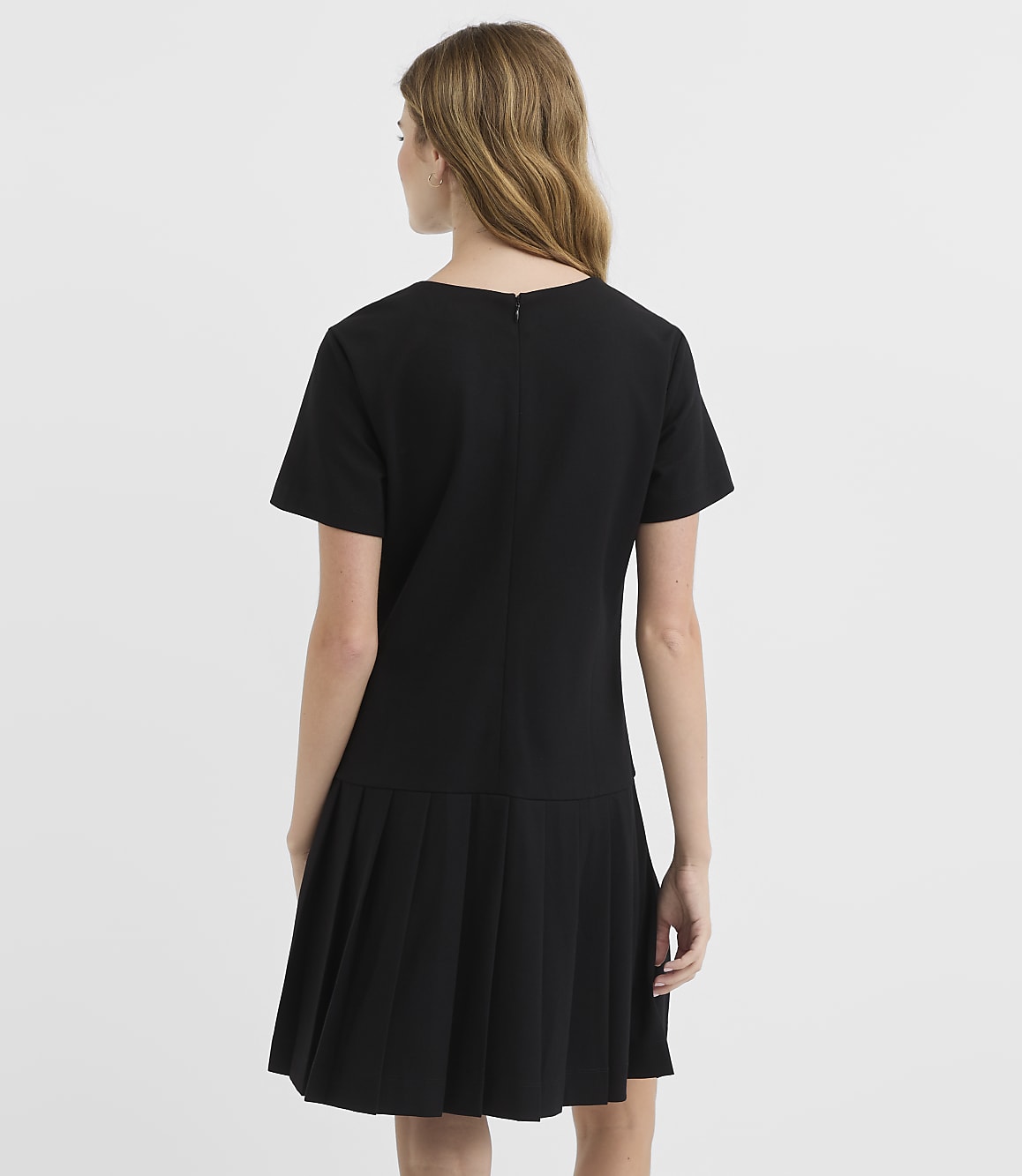 A woman in a black dress with a pleated skirt stands against a plain white background.