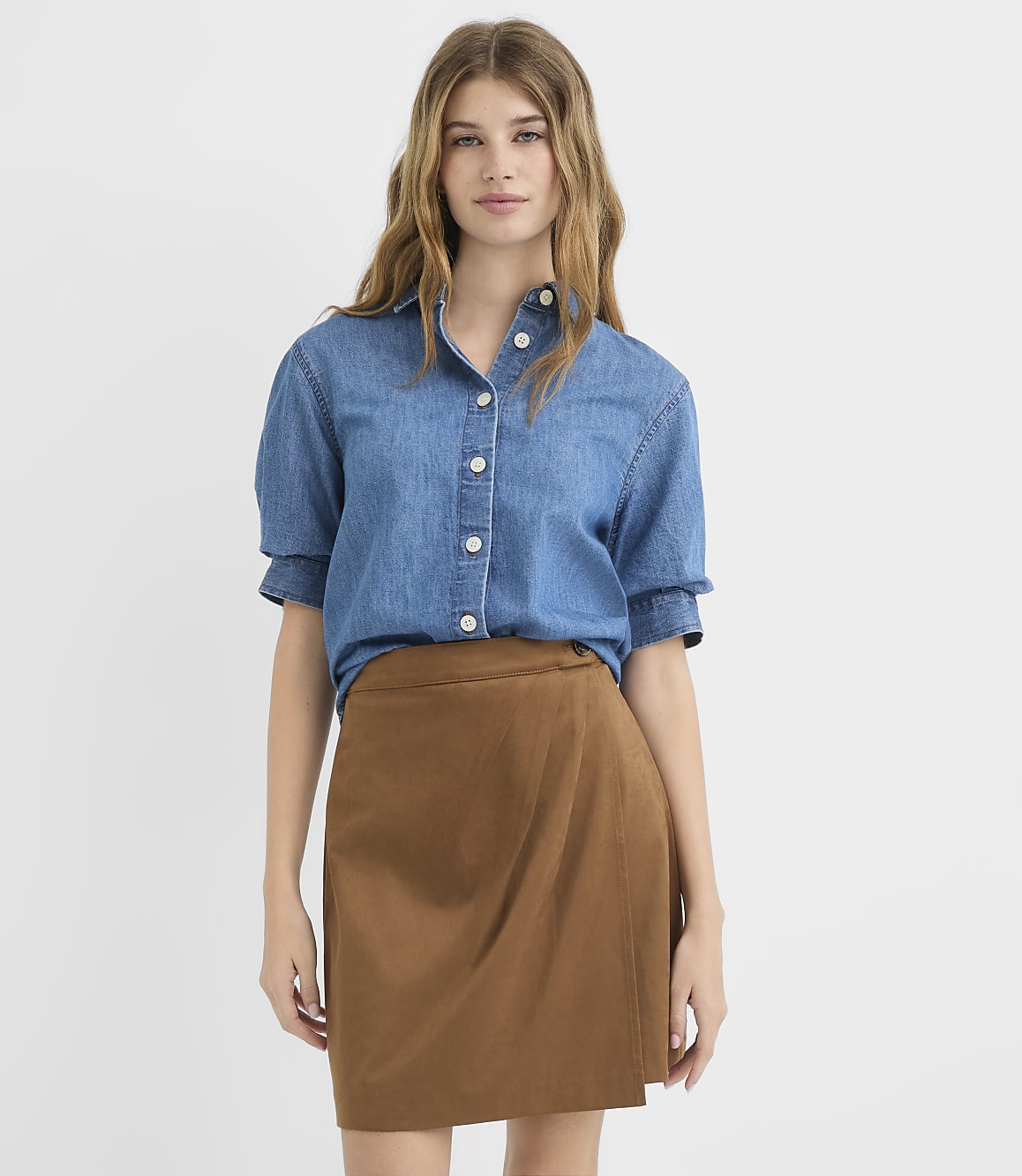 A young woman in a denim shirt and brown skirt stands against a plain white background.