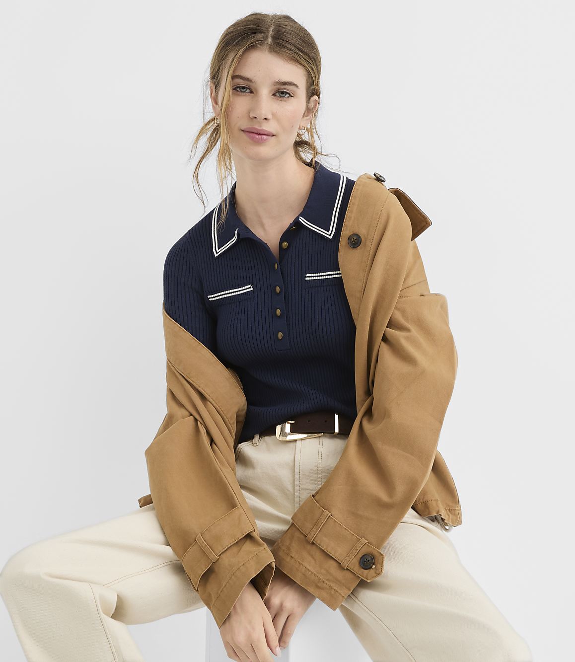 A woman in a navy polo shirt and tan jacket sits against a plain white background.