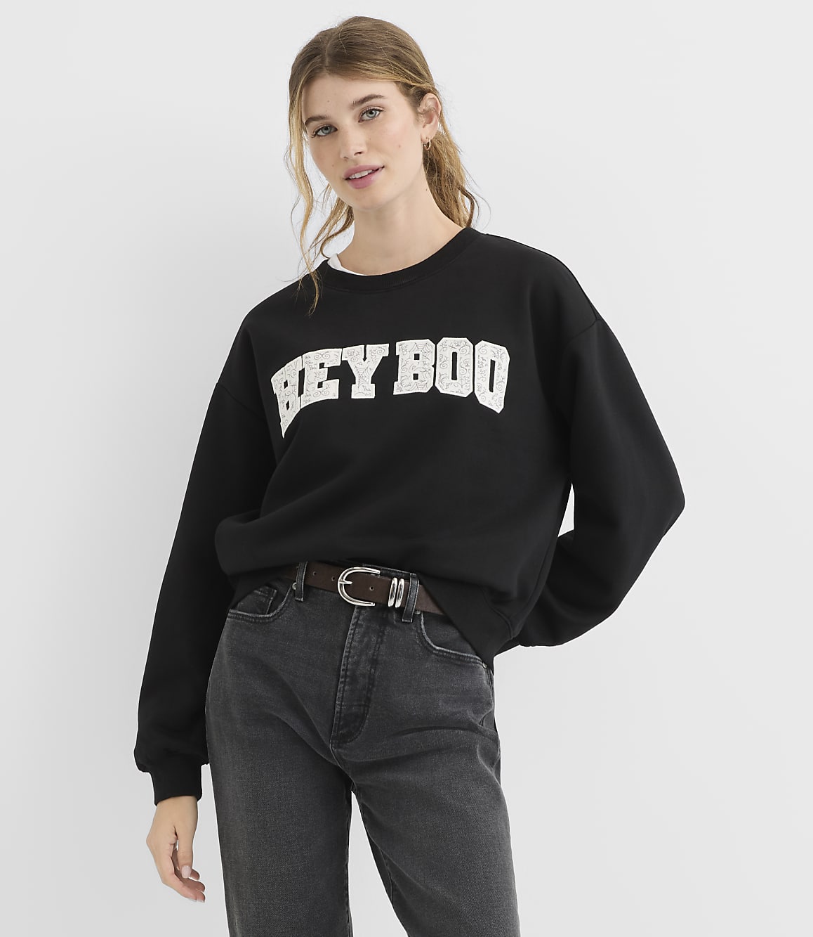 A young woman in a black sweatshirt with "HEY BOO" printed on it and dark jeans stands against a plain white background.