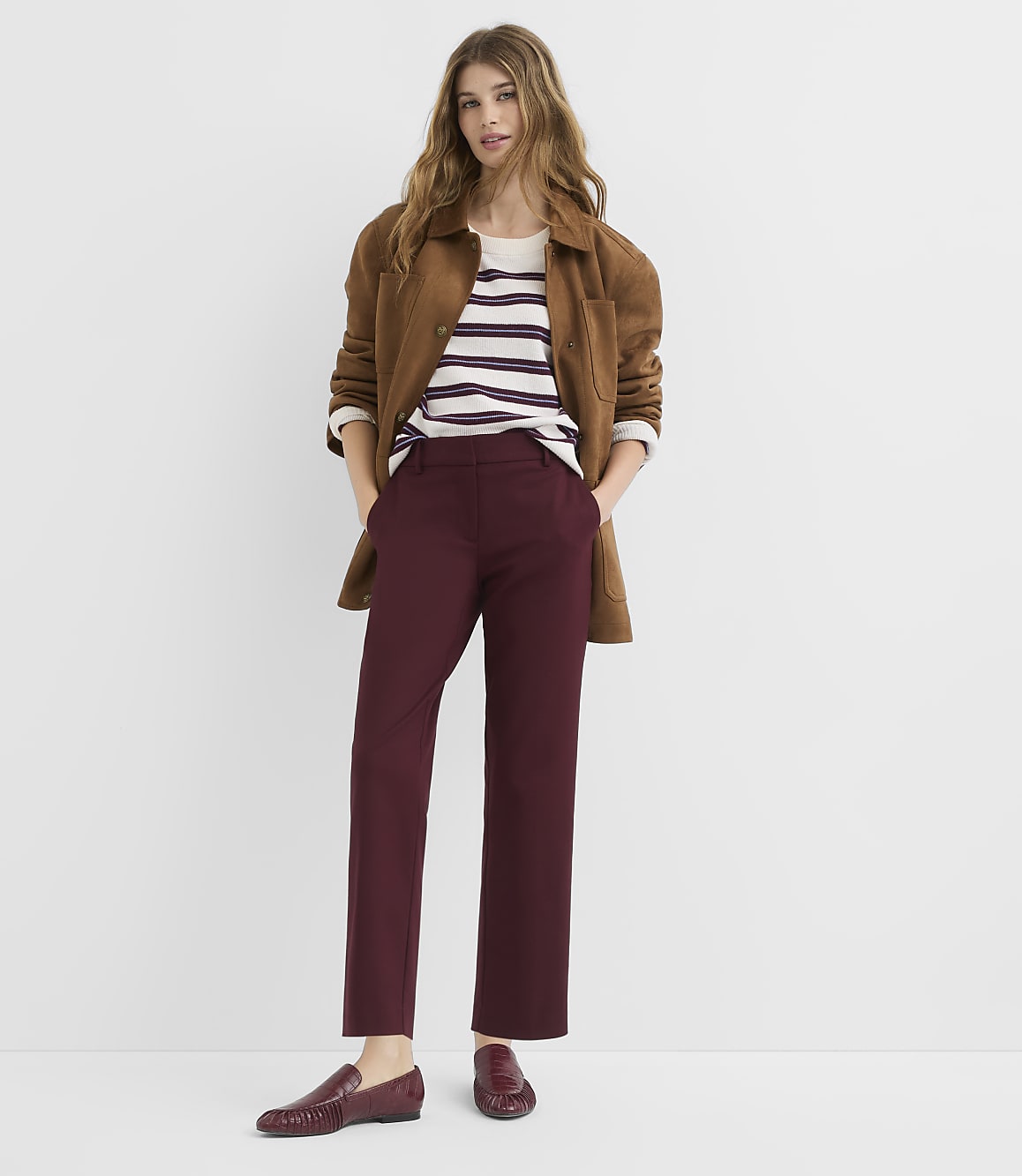 A woman in a brown jacket, striped shirt, and burgundy pants stands against a plain white background.