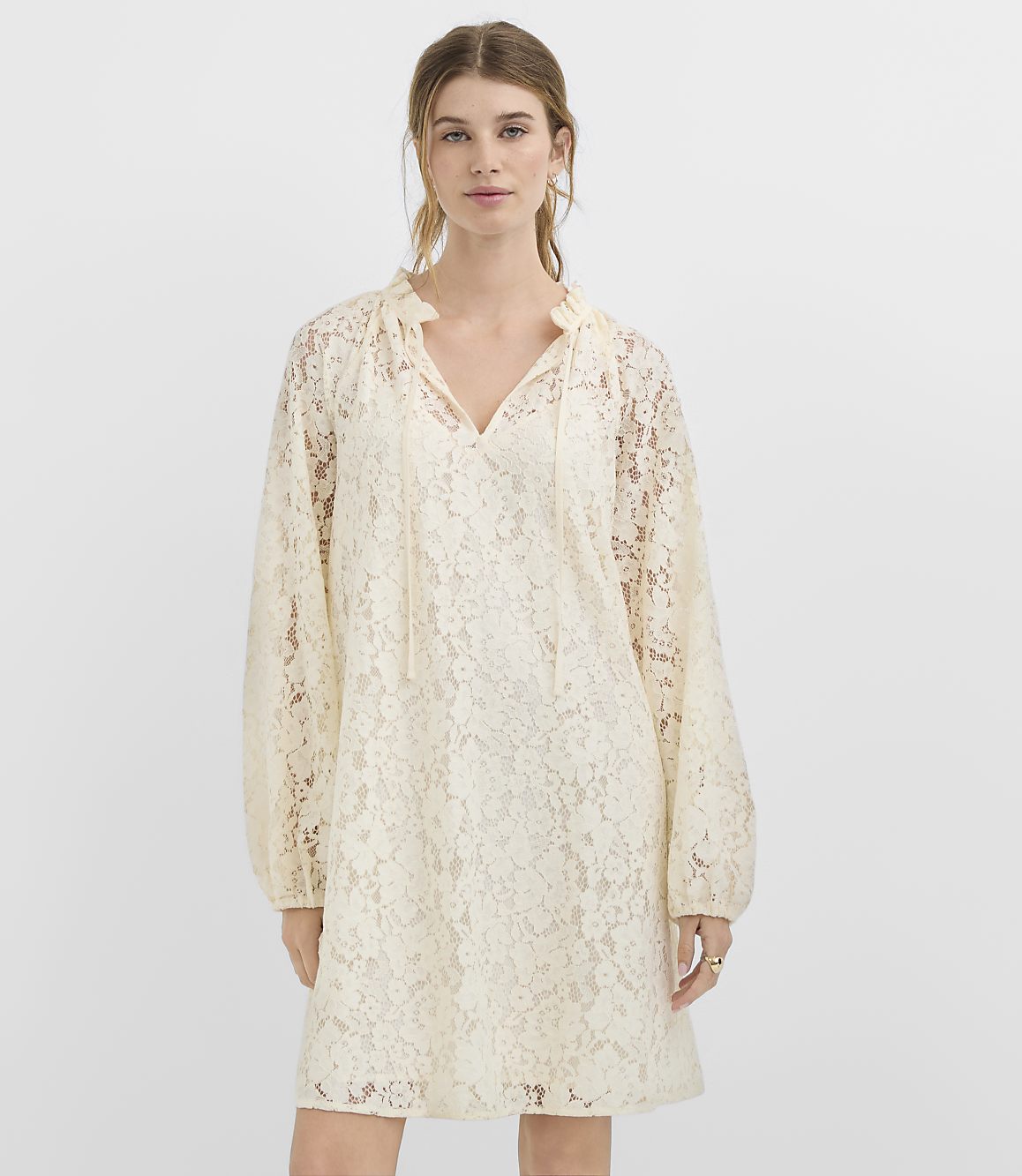 A woman in a cream lace dress stands against a plain white background.