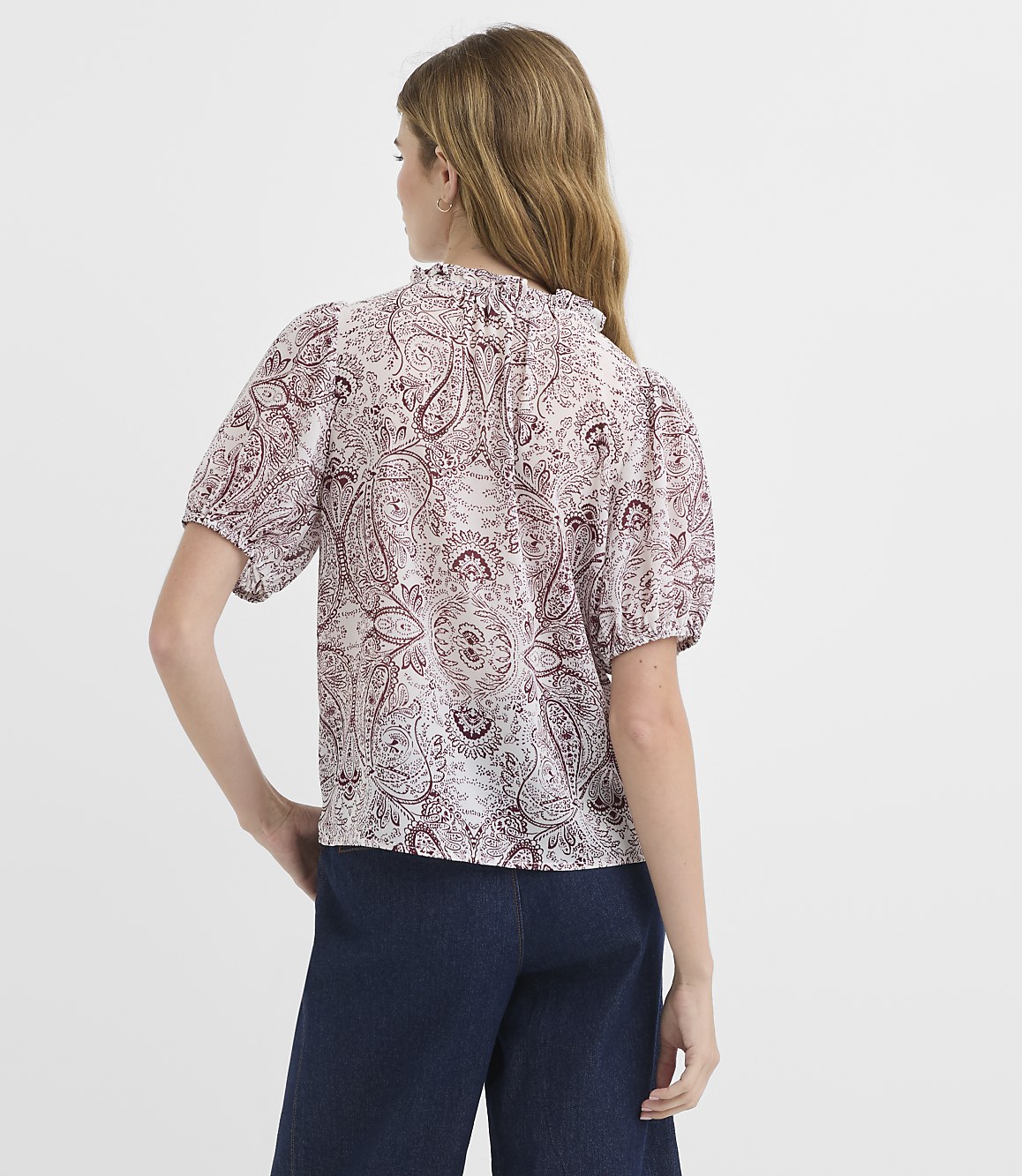 A woman in a paisley print blouse and dark jeans stands against a plain white background.