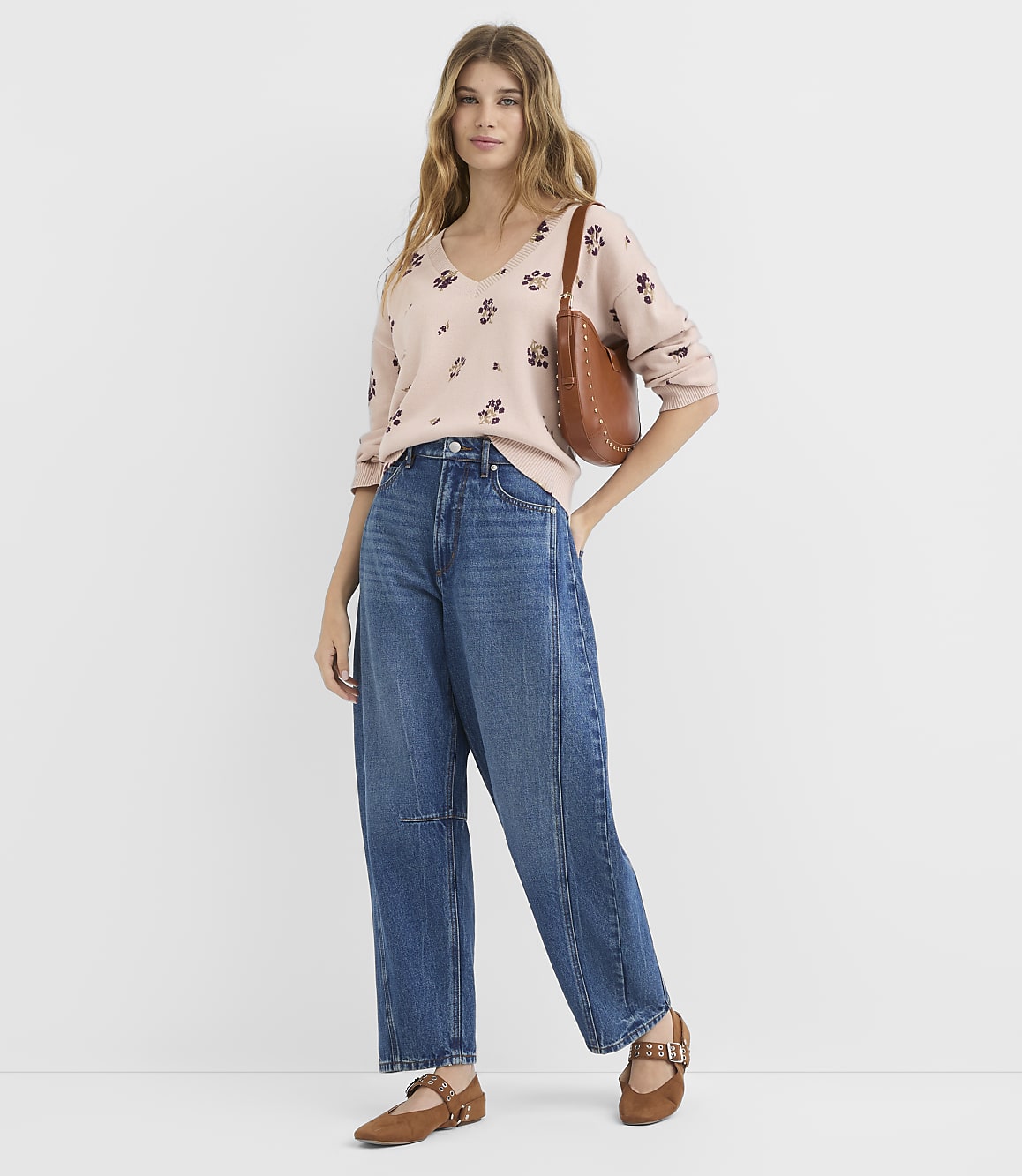 A woman in a floral sweater and wide-leg jeans stands against a plain white background.