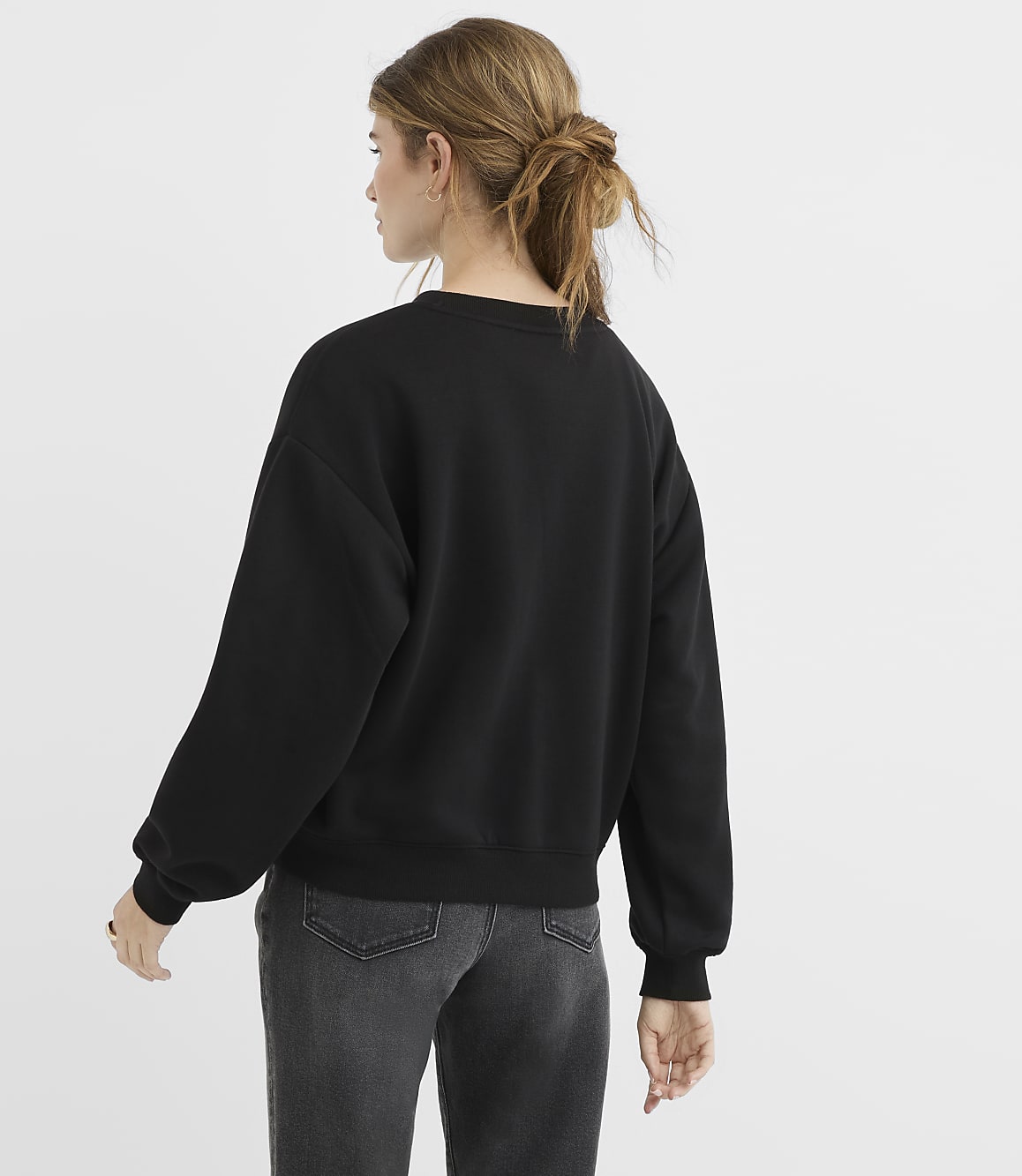 A woman in a black sweatshirt and grey jeans stands against a plain white background.