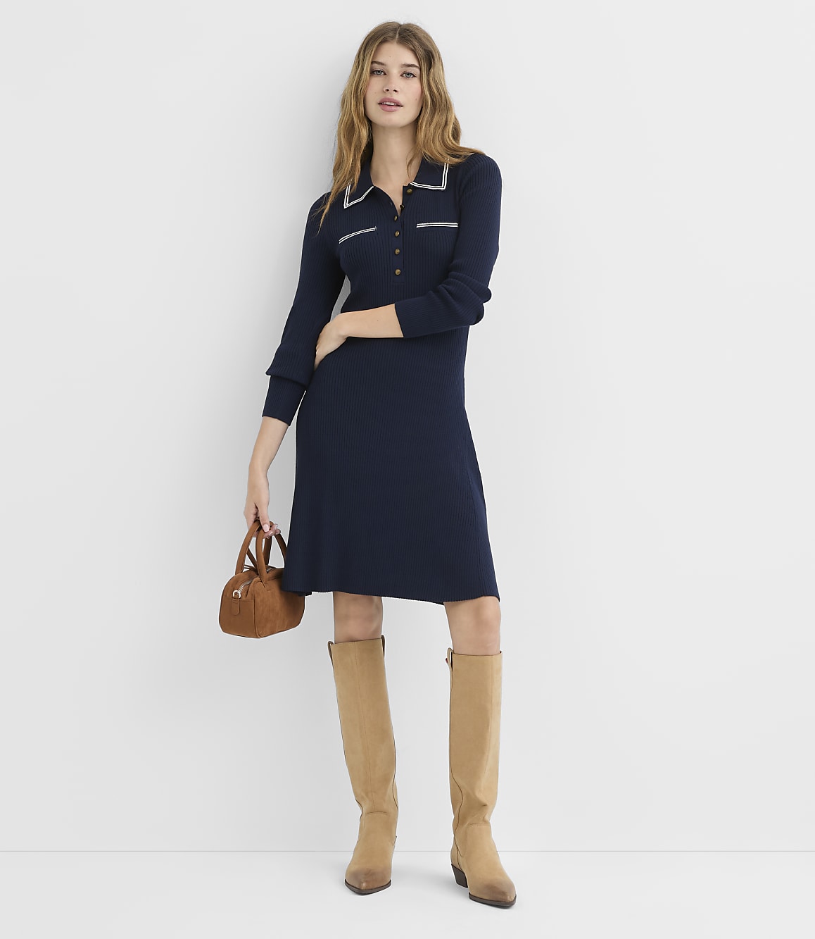 A woman in a navy blue dress and tan boots stands in front of a plain white background, holding a small brown purse.