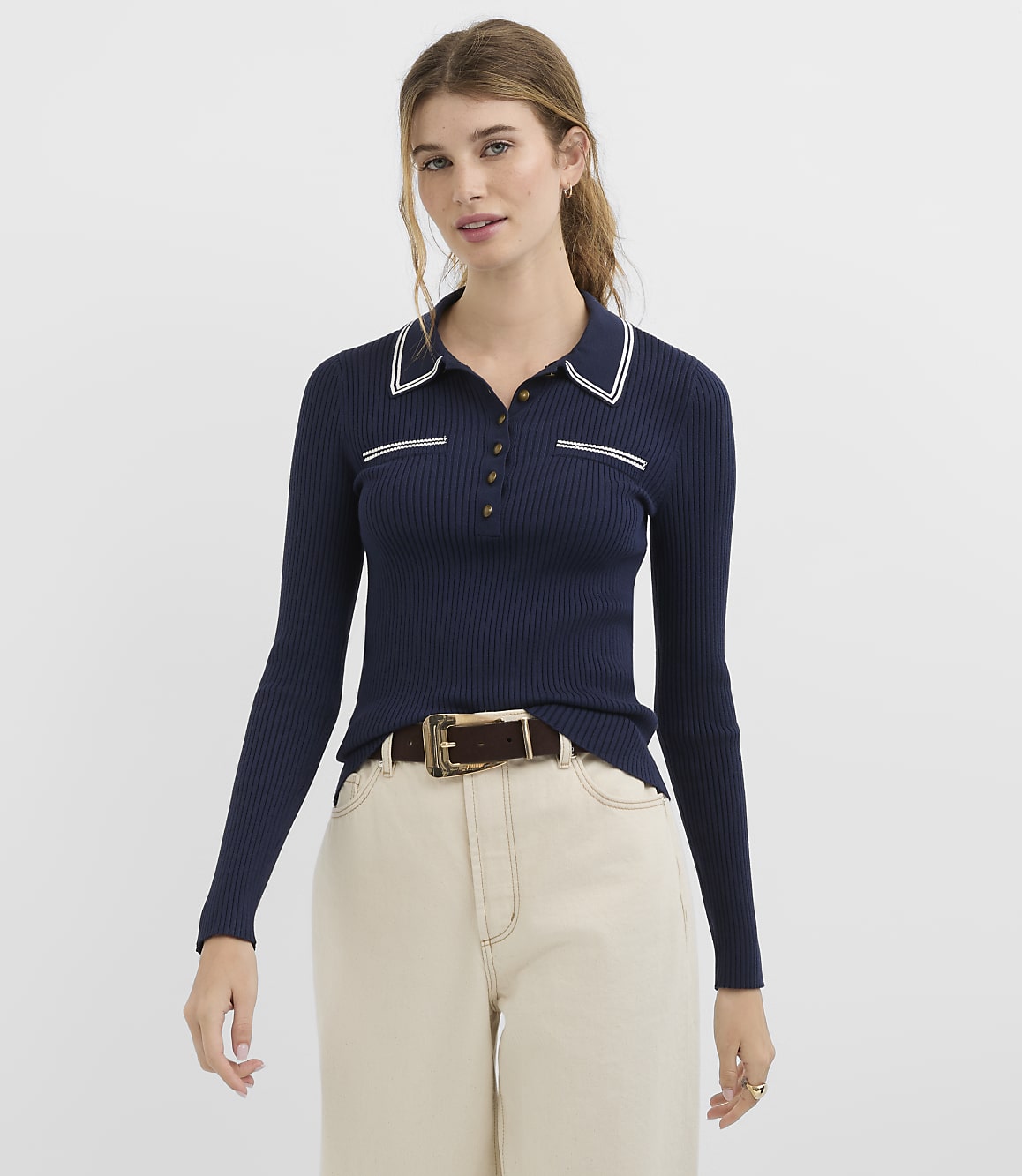 A woman in a navy ribbed polo shirt and cream pants stands against a plain white background.