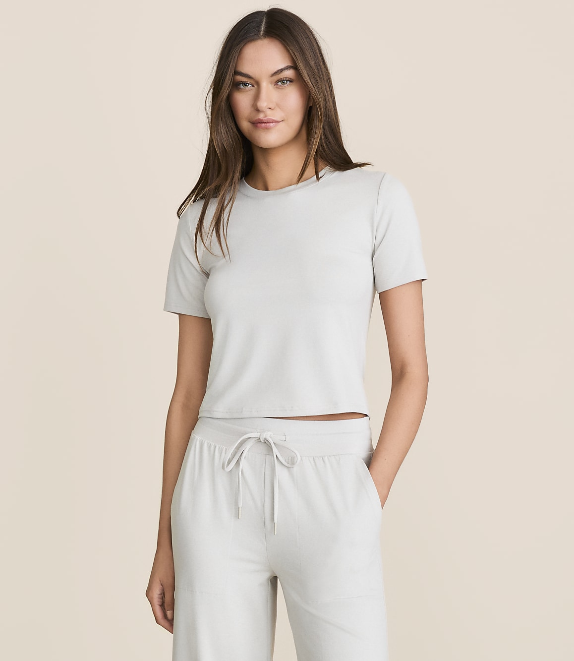 A woman in a light gray cropped t-shirt and matching drawstring pants stands against a plain, light beige background.