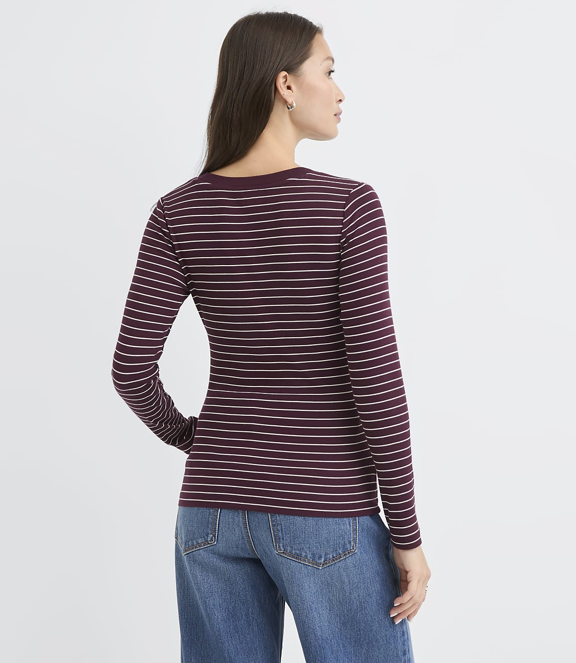 A woman in a striped long-sleeved shirt and jeans stands against a plain white background.