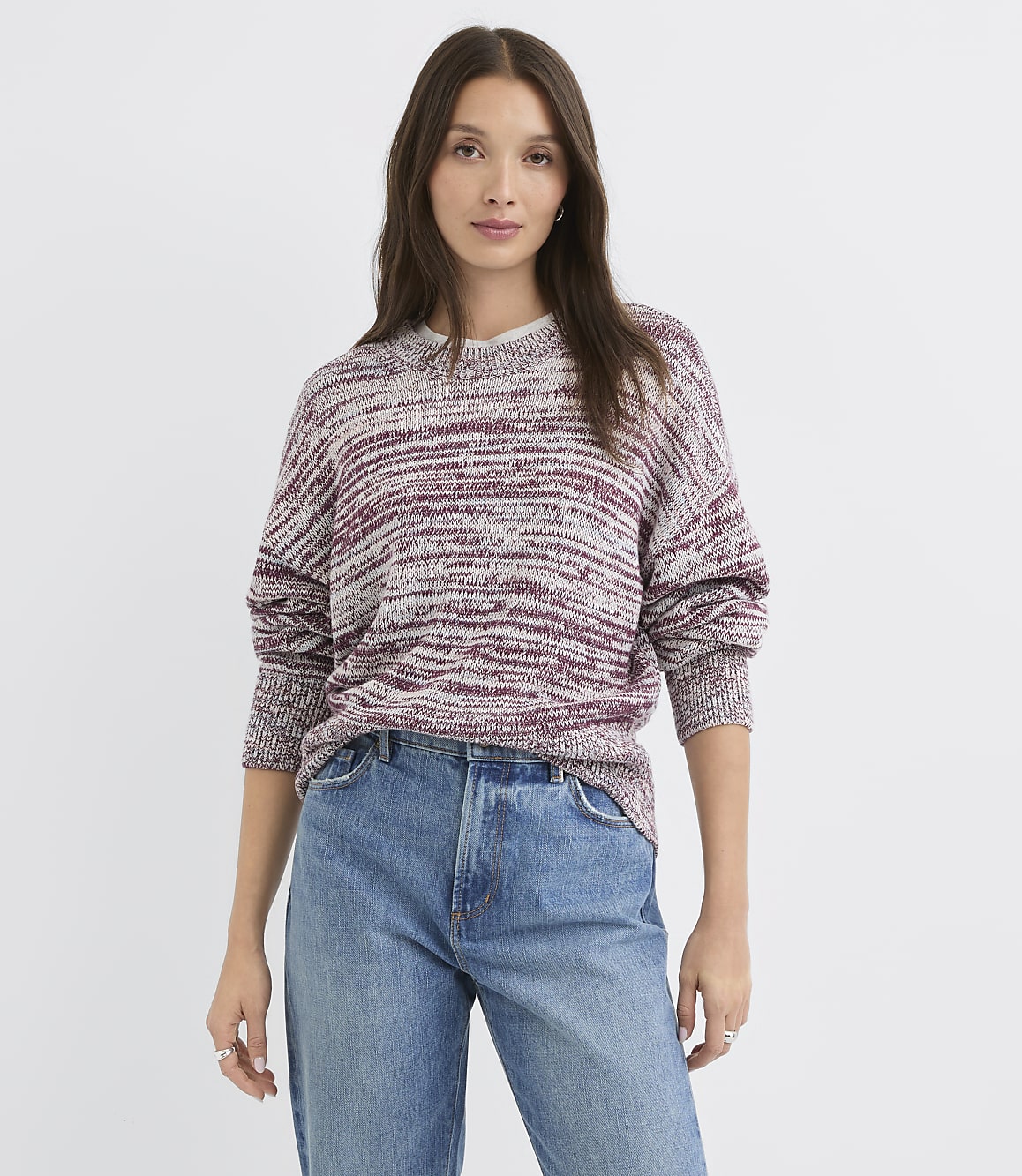 A woman in a marled burgundy and grey sweater and blue jeans stands against a plain white background.