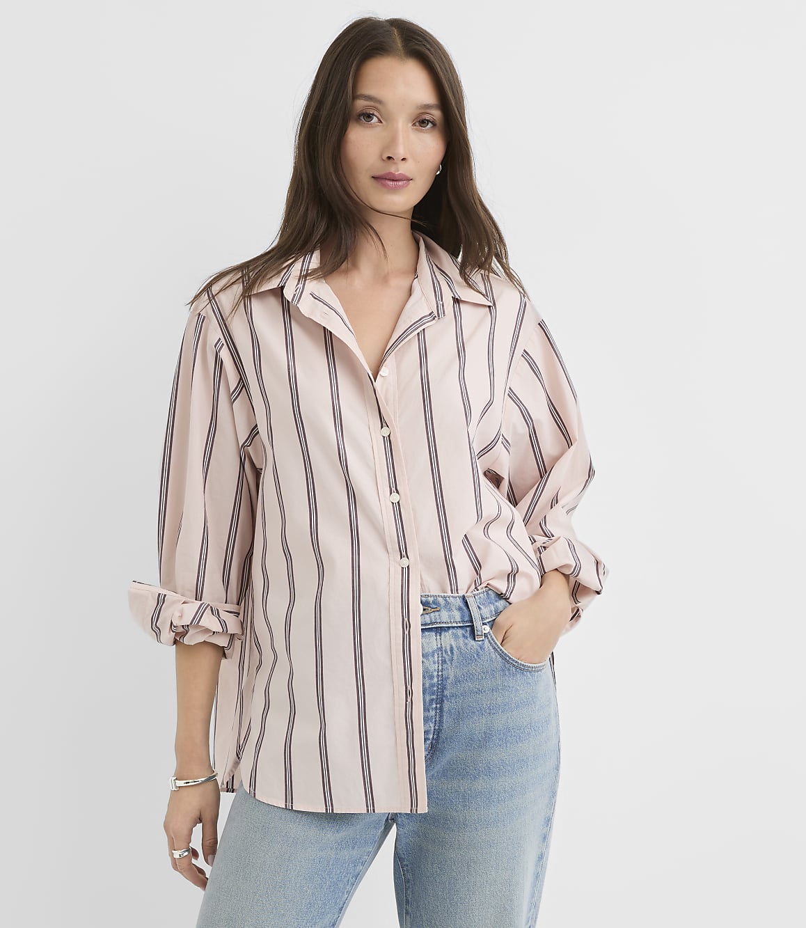 A woman in a pink and black striped button-down shirt and light blue jeans stands against a plain white background.