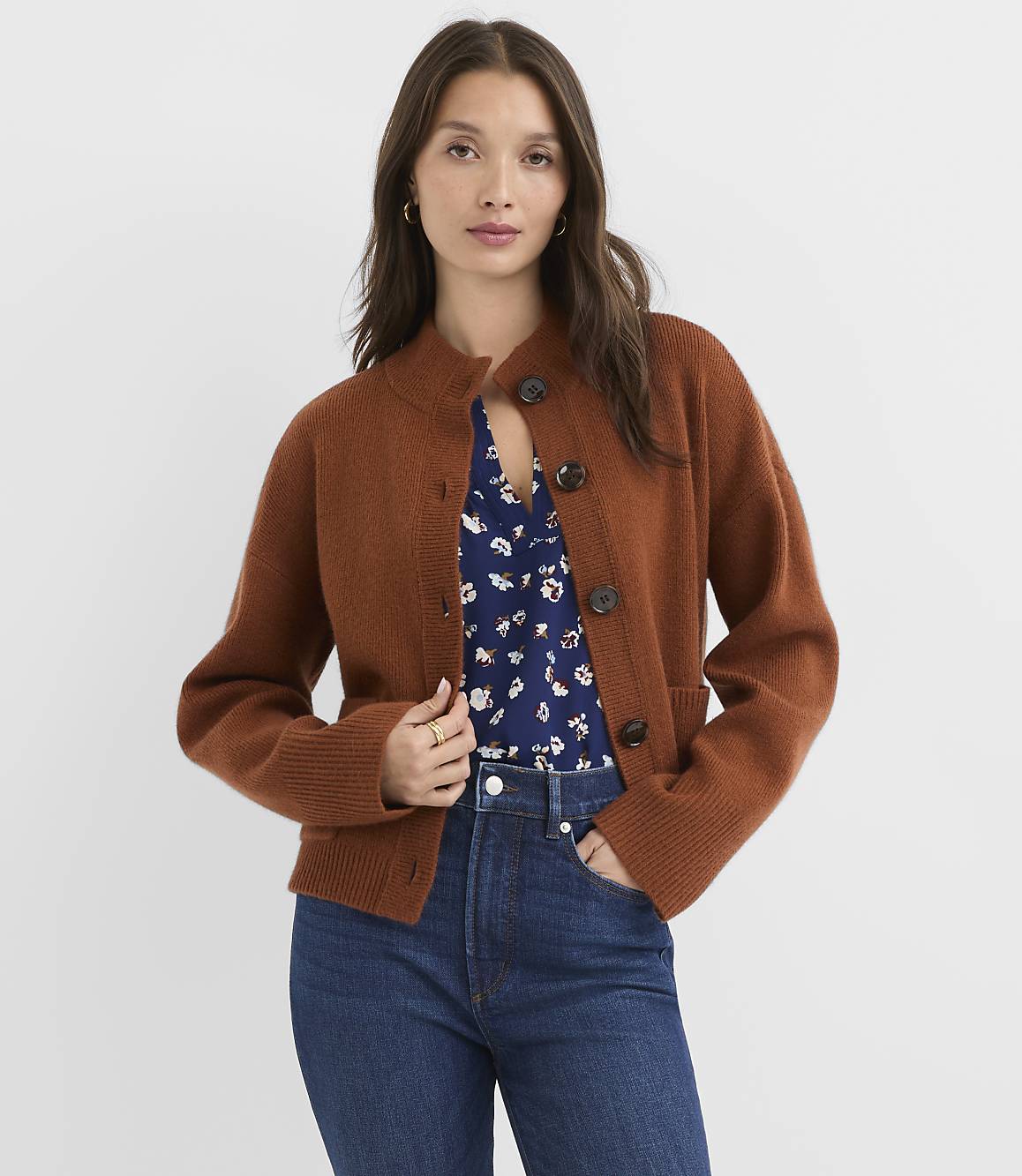 A woman in a brown cardigan and blue floral top stands against a plain white background.