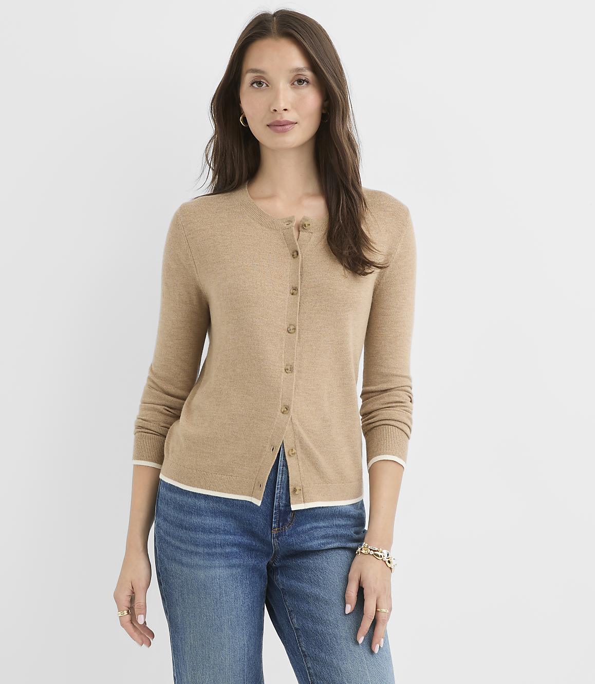 A woman in a tan cardigan and blue jeans stands against a plain white background.