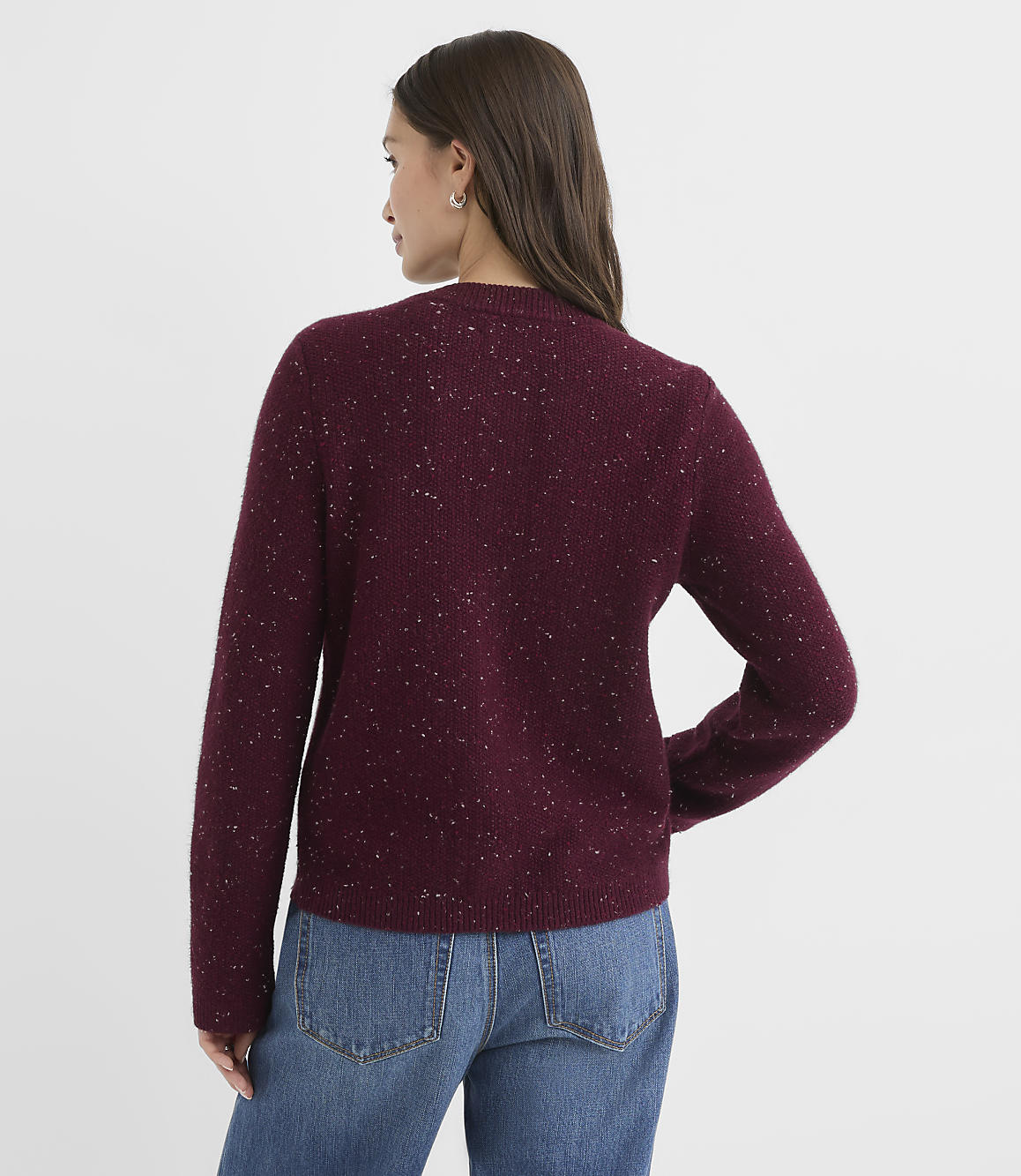 A woman in a maroon speckled sweater and blue jeans stands against a plain white background.