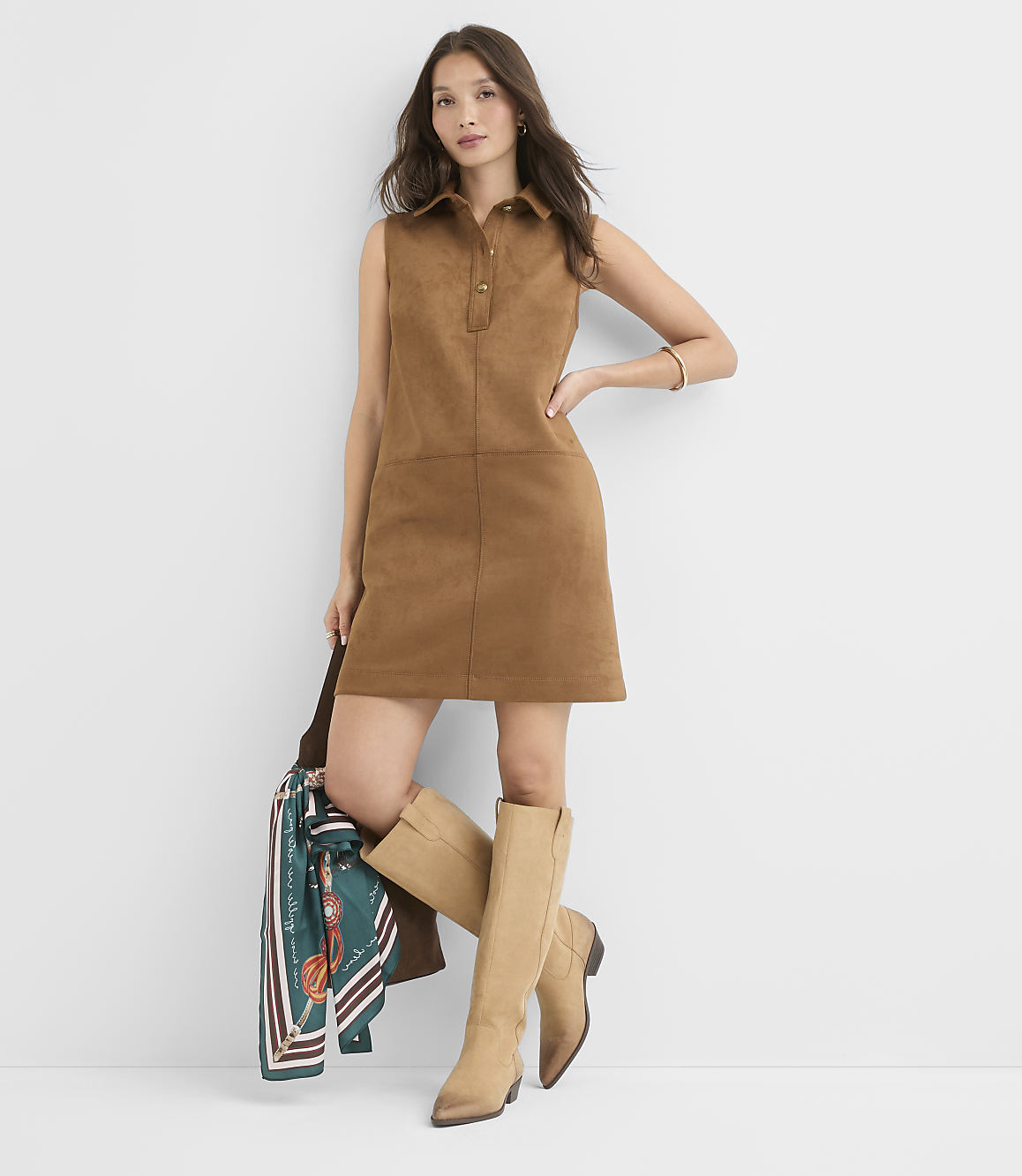A woman in a brown suede dress and tan boots poses against a plain white background, holding a patterned scarf.