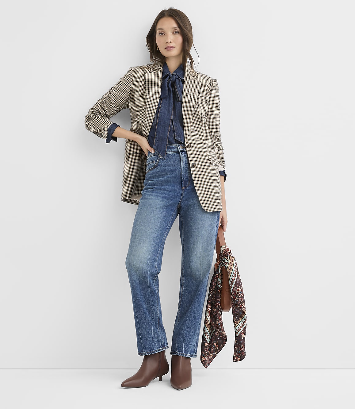 A woman in a patterned blazer, denim shirt, and jeans stands against a plain white background, holding a patterned scarf.