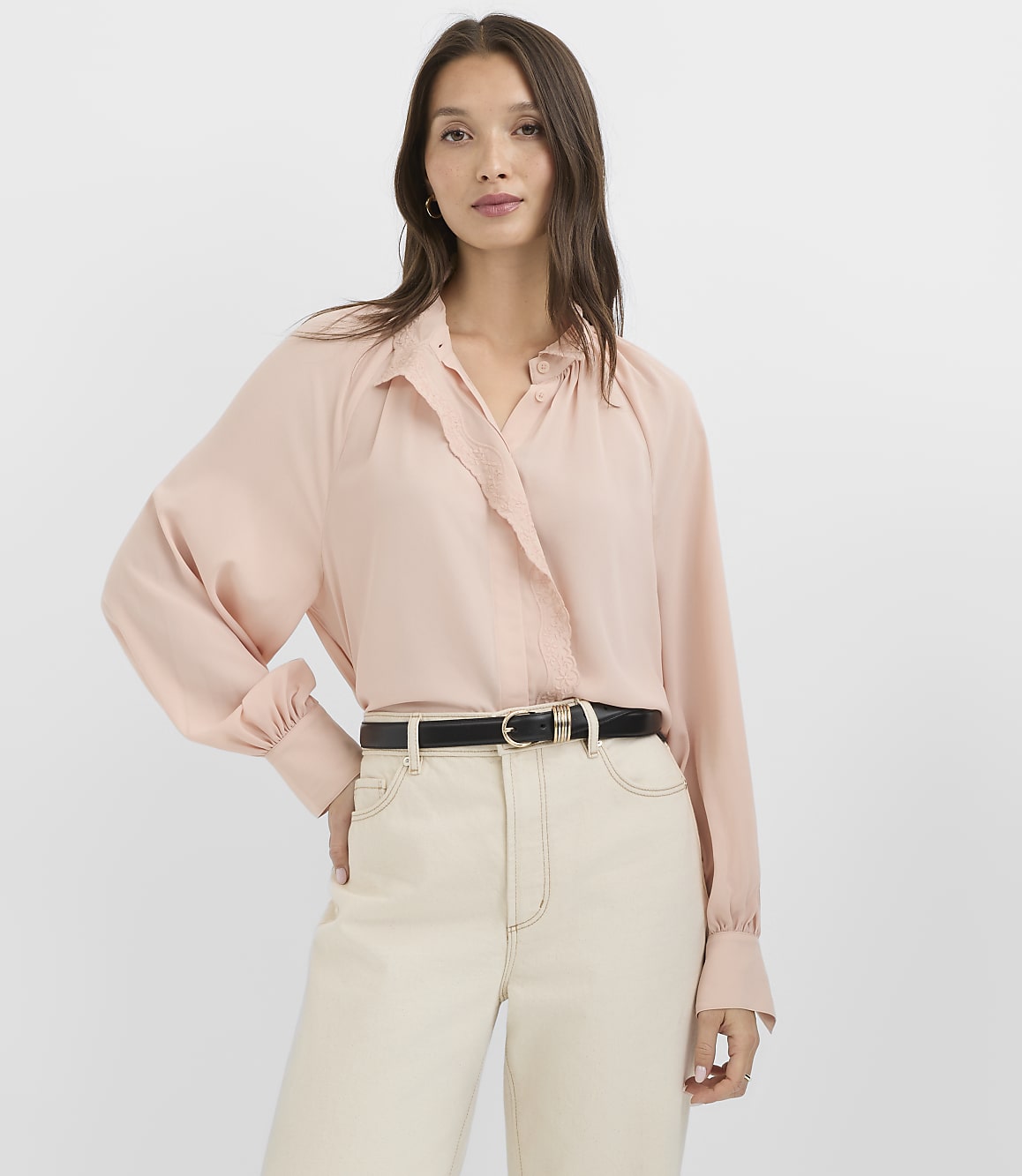 A woman in a pale pink blouse and cream pants stands against a plain white background.