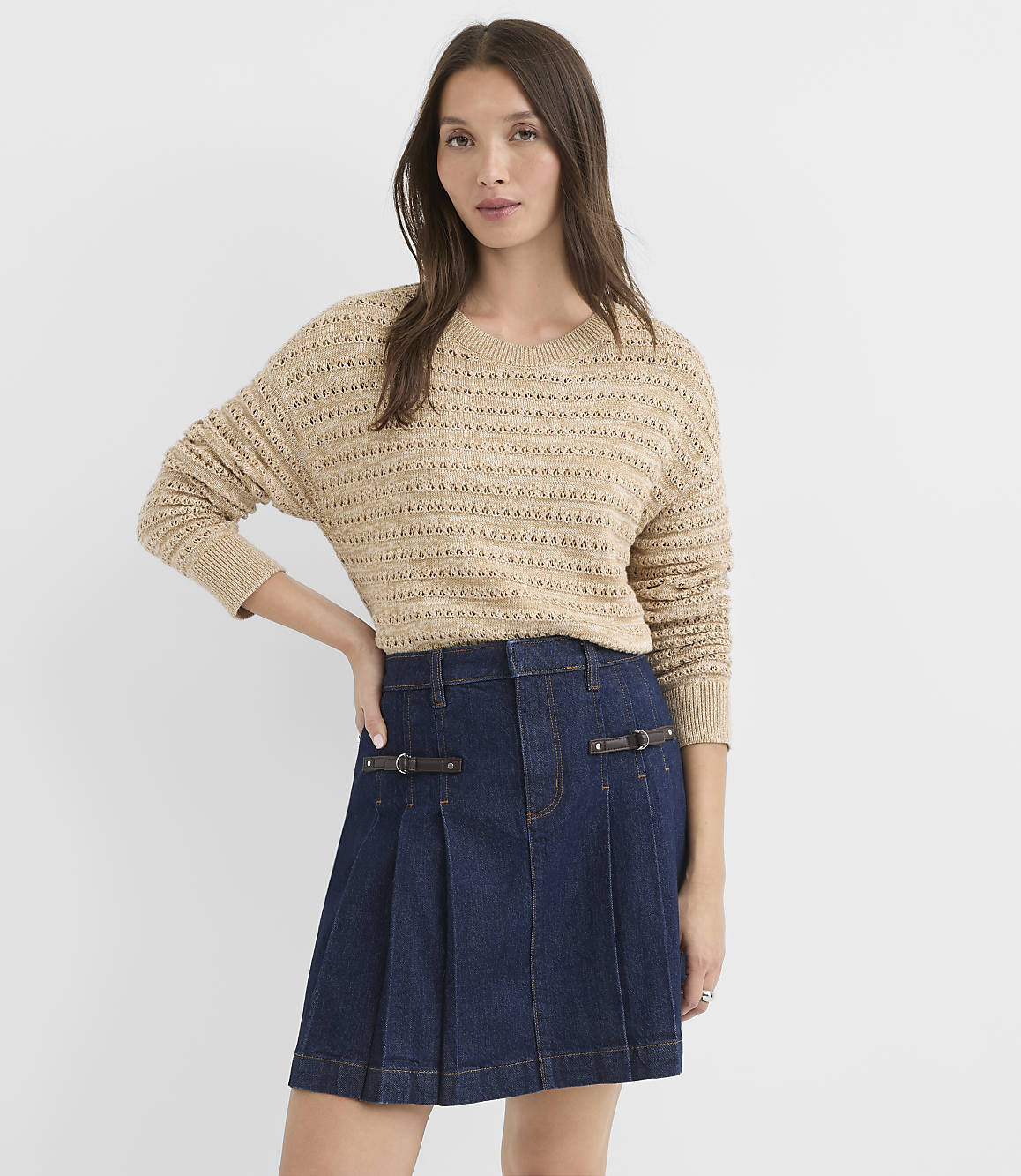 A woman in a tan knit sweater and a dark denim skirt stands against a plain white background.