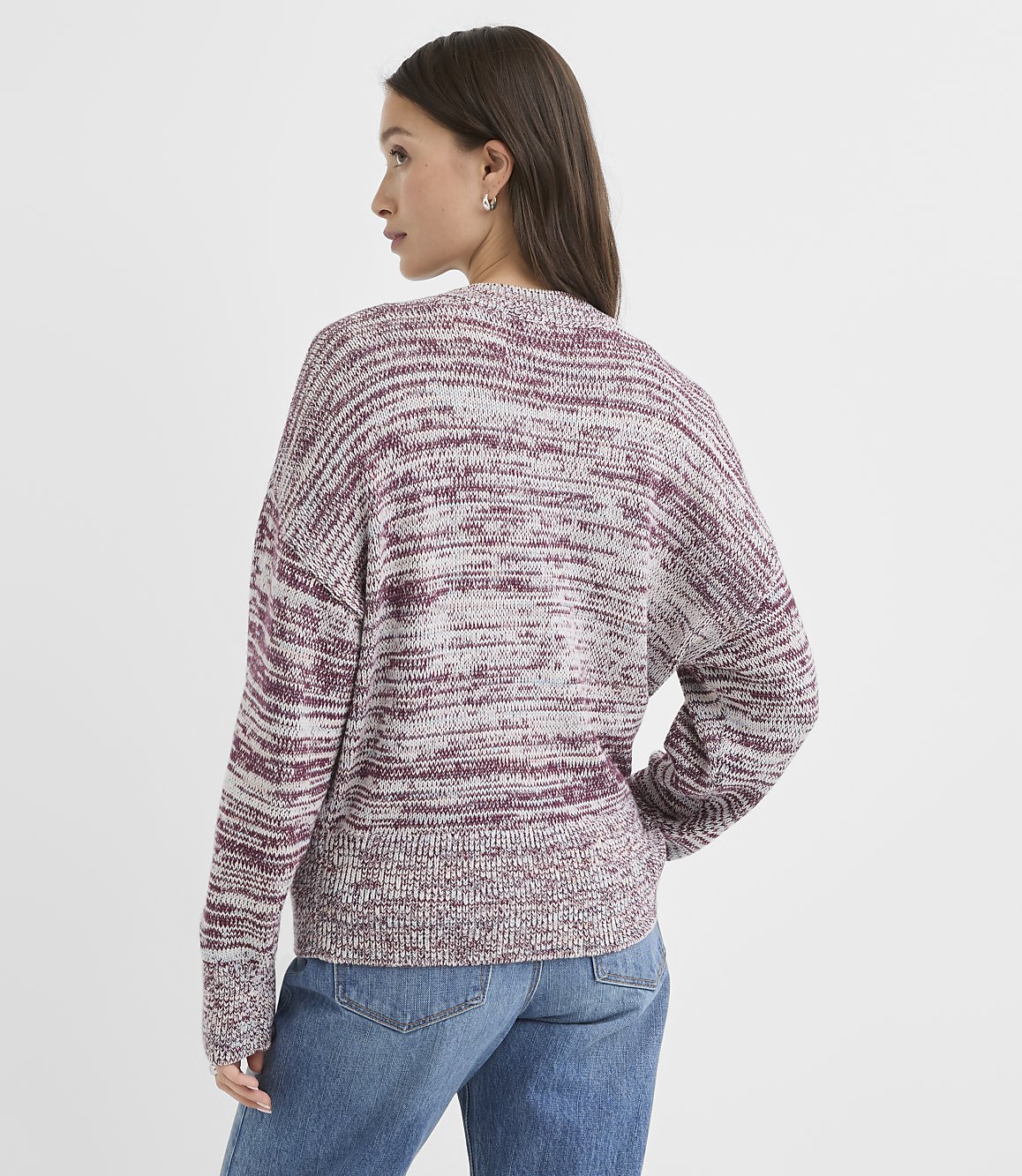 A woman in a marled burgundy and grey sweater and blue jeans stands against a plain white background.
