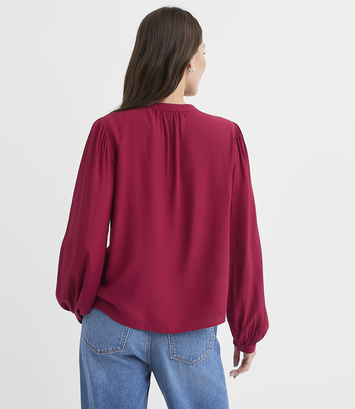 A woman in a red blouse and blue jeans stands against a plain white background.