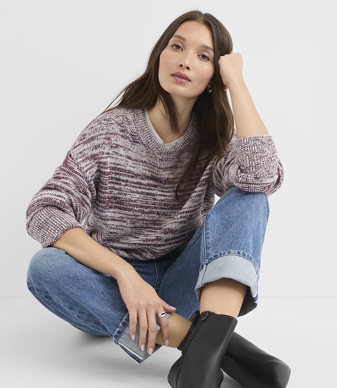 A woman in a marled burgundy and white sweater and cuffed blue jeans sits on a white background, leaning her head on her hand.