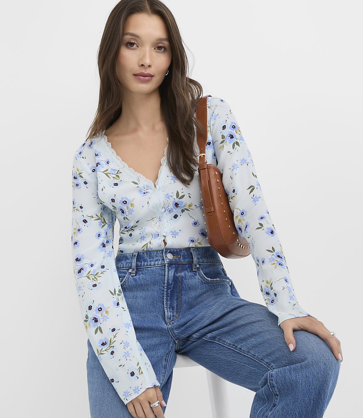A woman in a light blue floral cardigan and blue jeans poses with a brown shoulder bag against a plain white background.