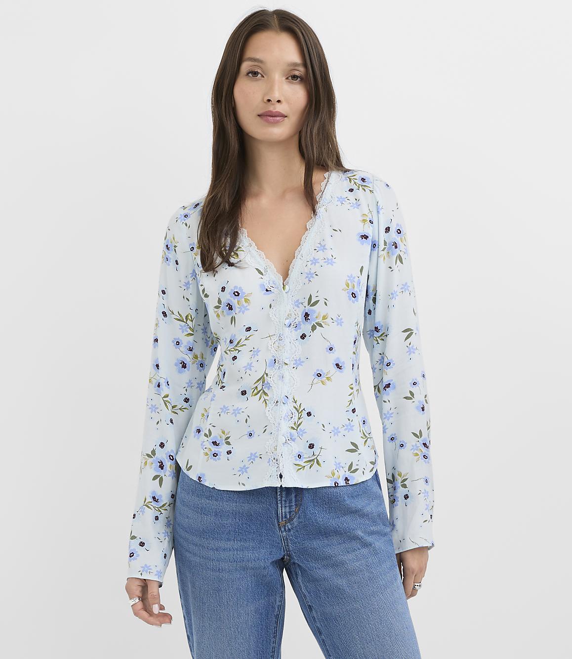 A woman in a light blue floral blouse and jeans stands against a plain white background.