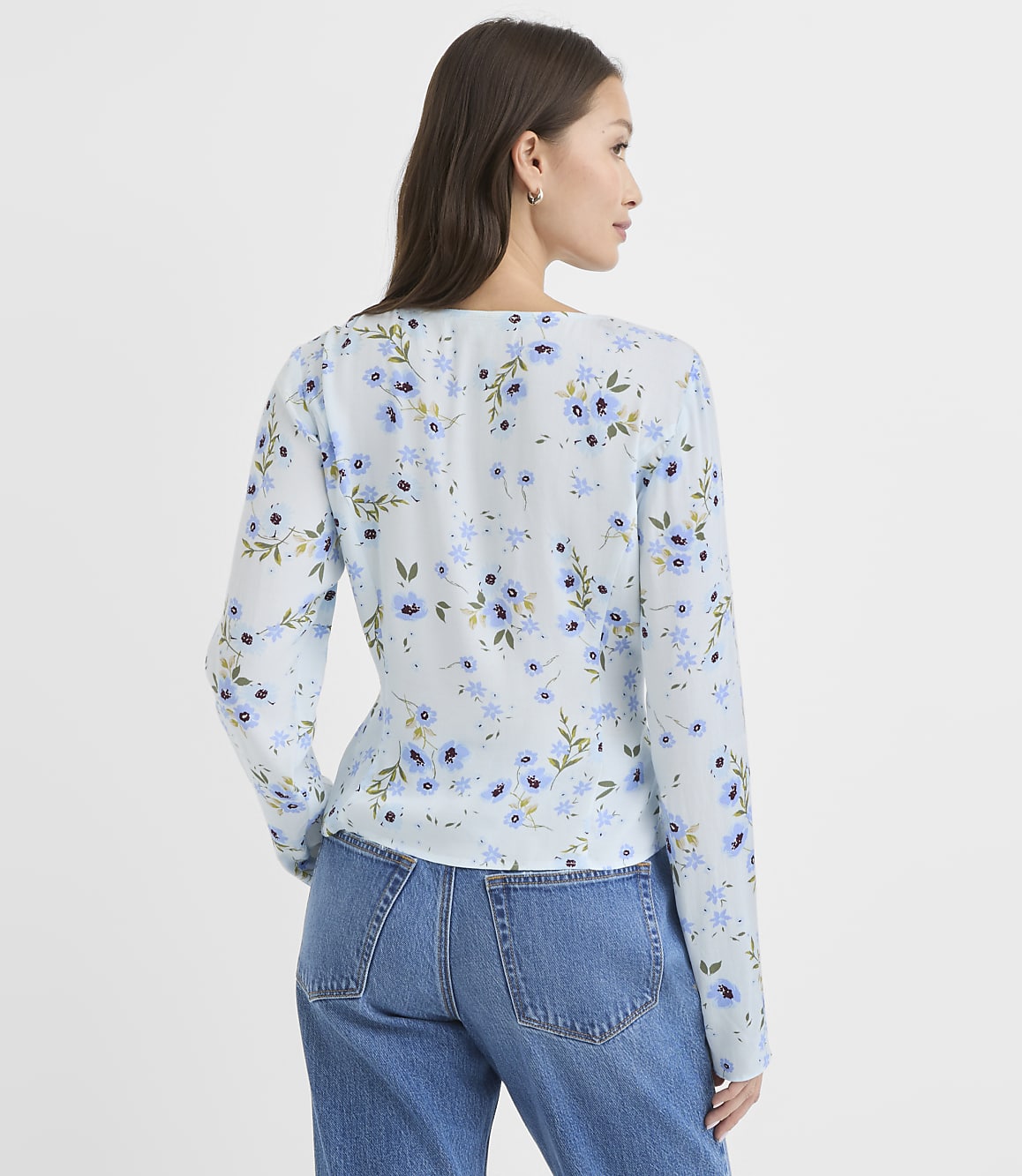 A woman in a light blue floral blouse and jeans stands against a plain white background.