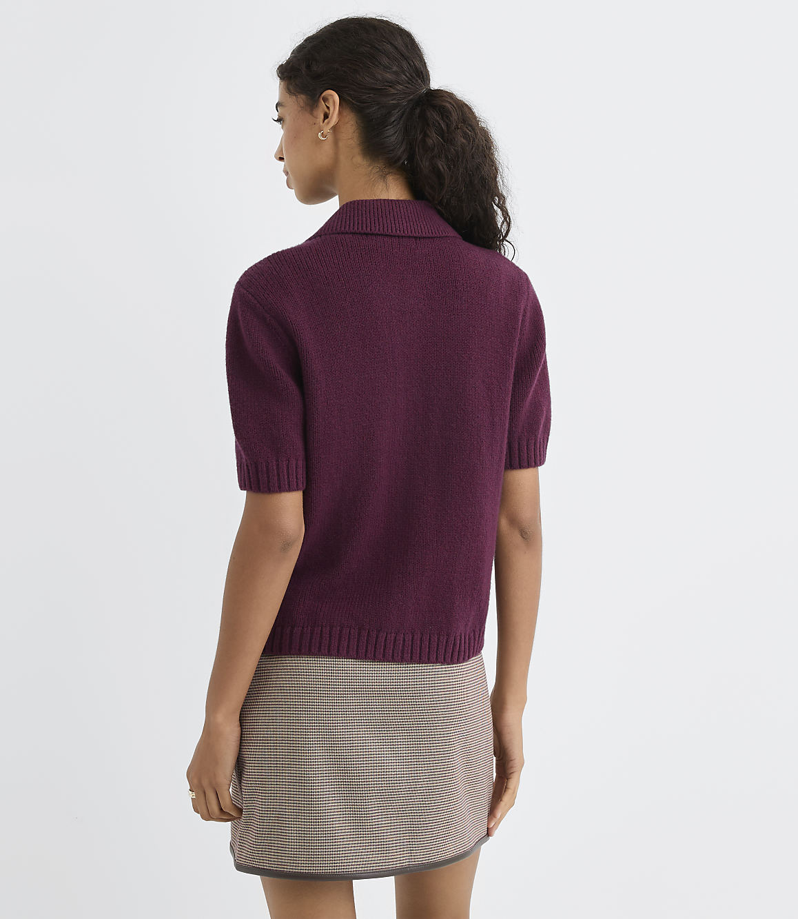 A woman in a burgundy collared sweater and a plaid skirt stands against a plain white background.