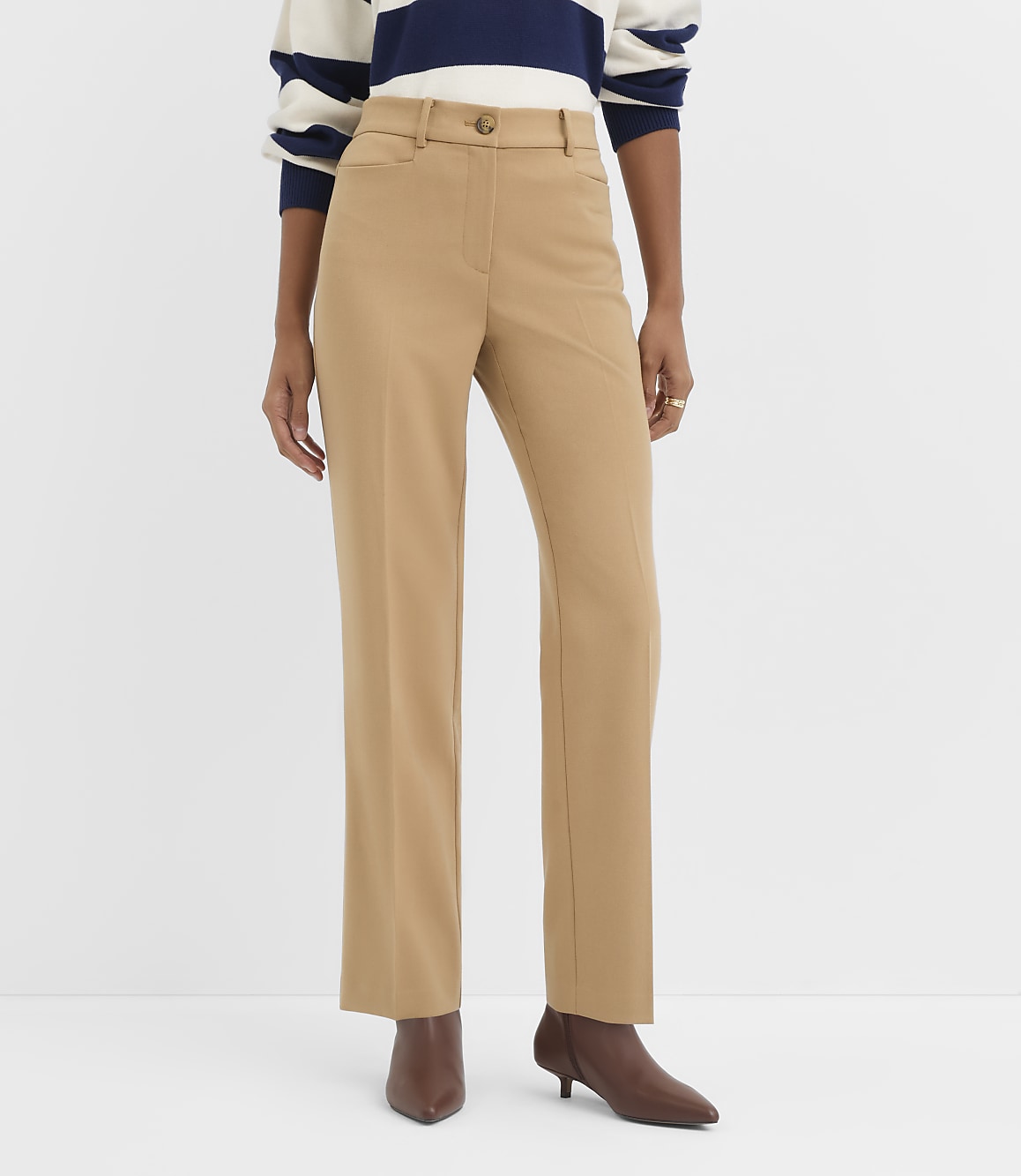 A person stands in the foreground wearing tan trousers and brown heeled boots, with a plain white background behind them.