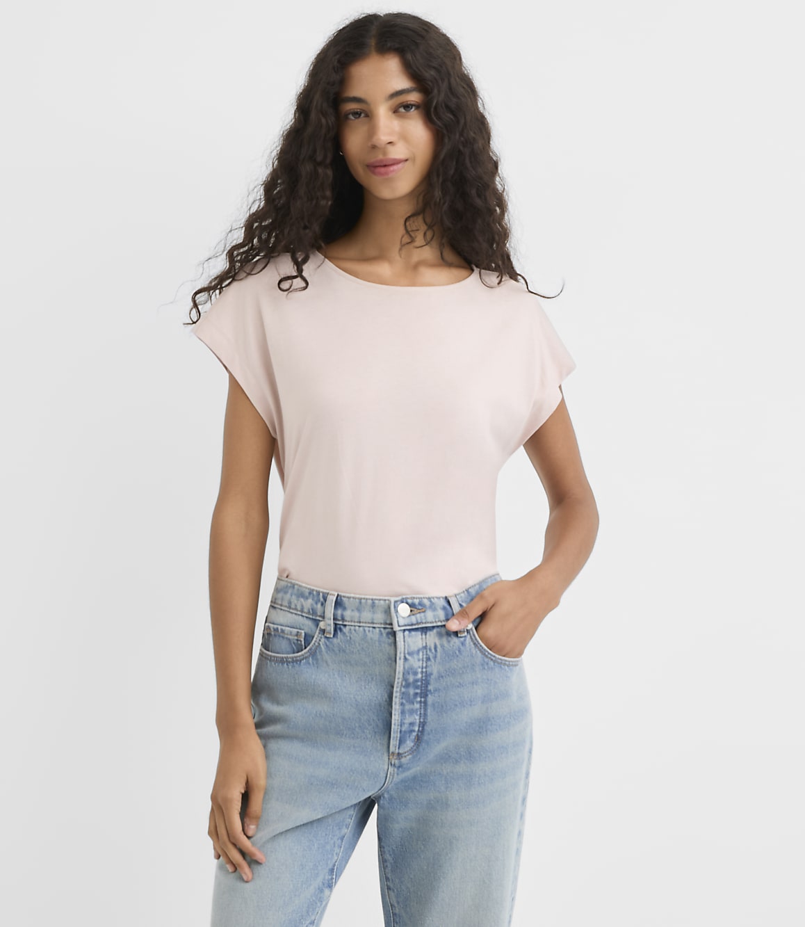 A woman in a light pink t-shirt and blue jeans stands against a plain white background.