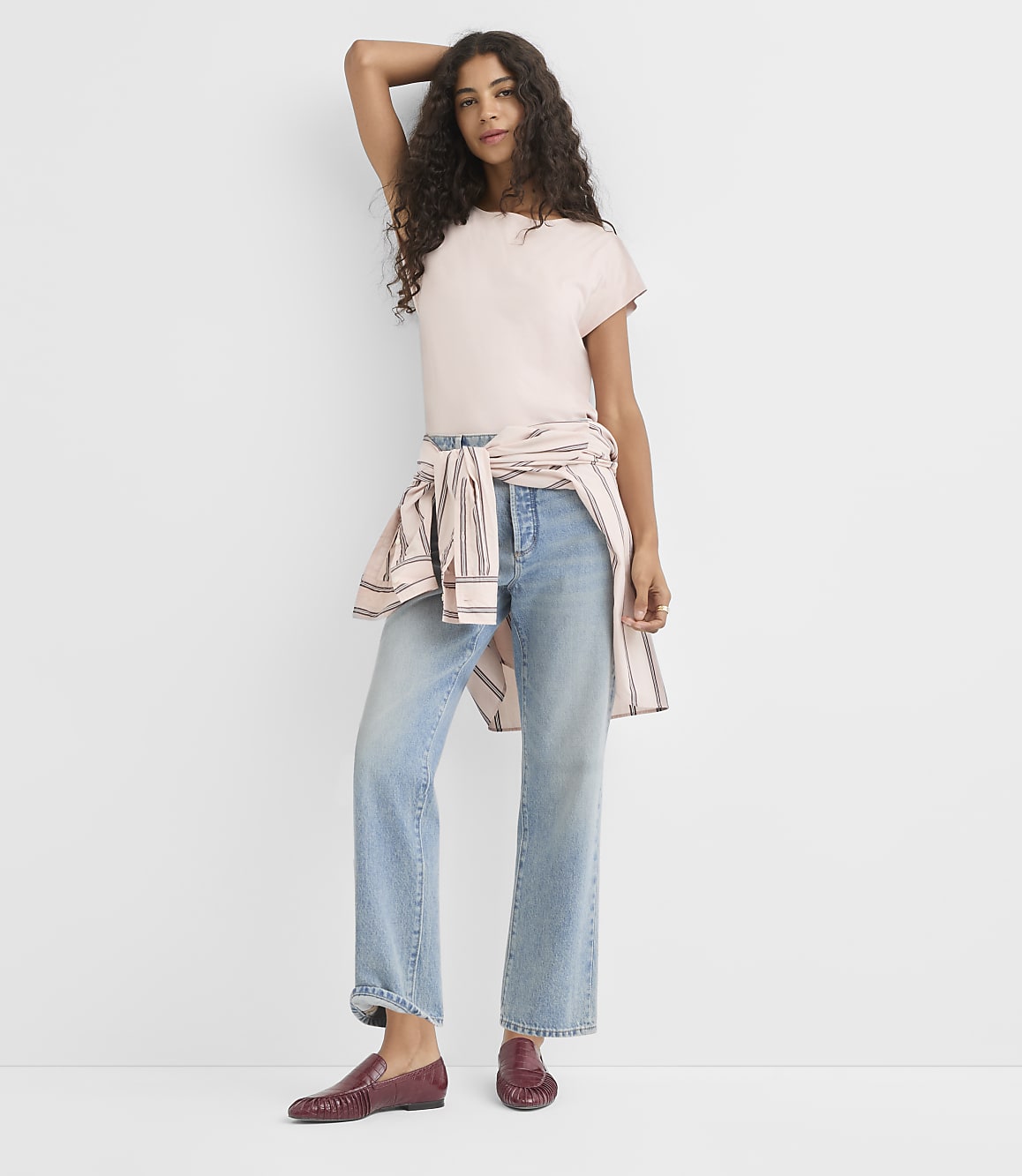A woman in a pink t-shirt and light wash jeans stands against a plain white background.